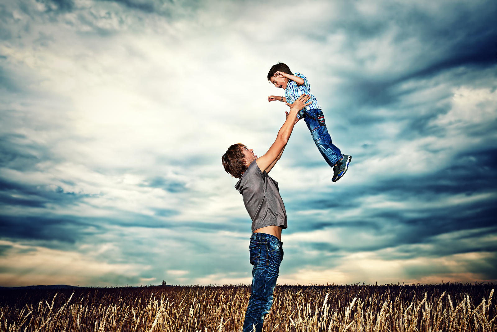 A man stands in a wheat field lifting a young boy into the air, both smiling under a dramatic, cloudy sky.
