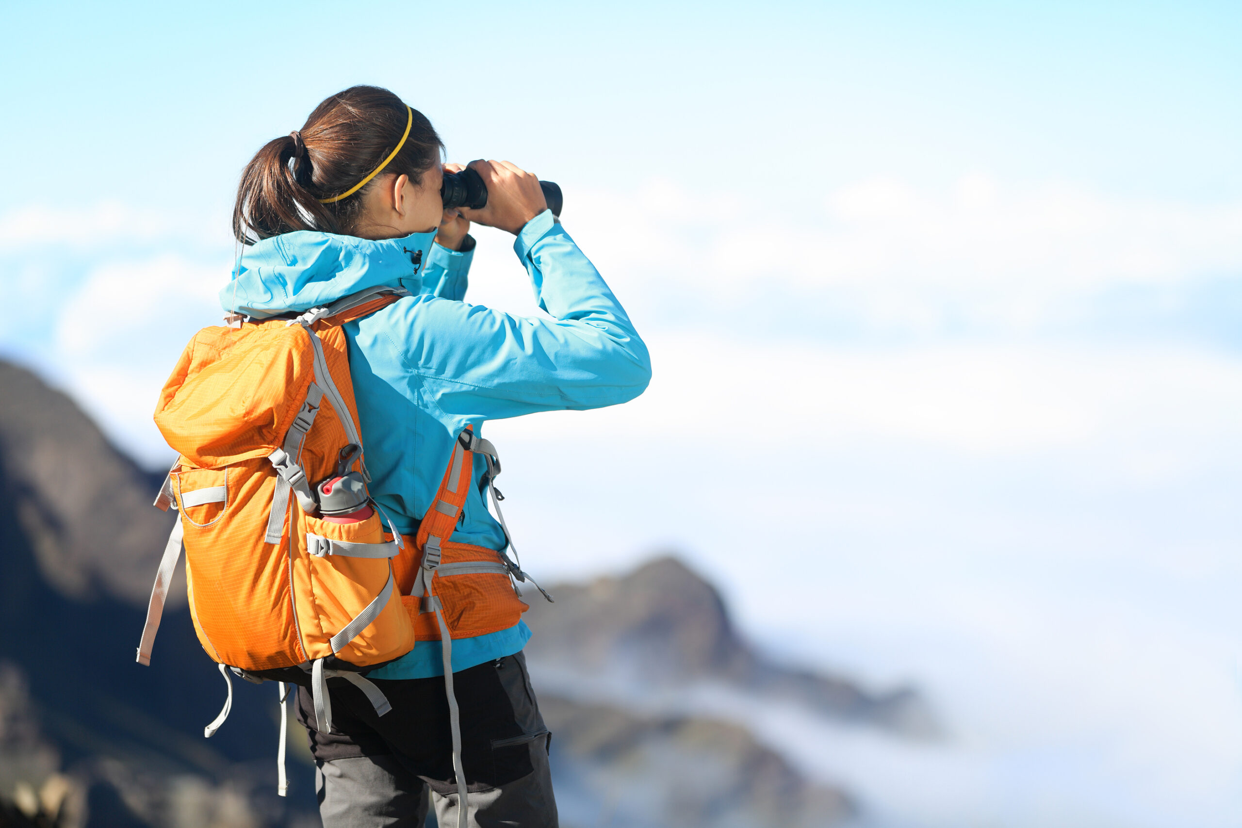 A person wearing a blue jacket and orange backpack looks through binoculars while standing on a mountain with a scenic, cloudy landscape in the background.