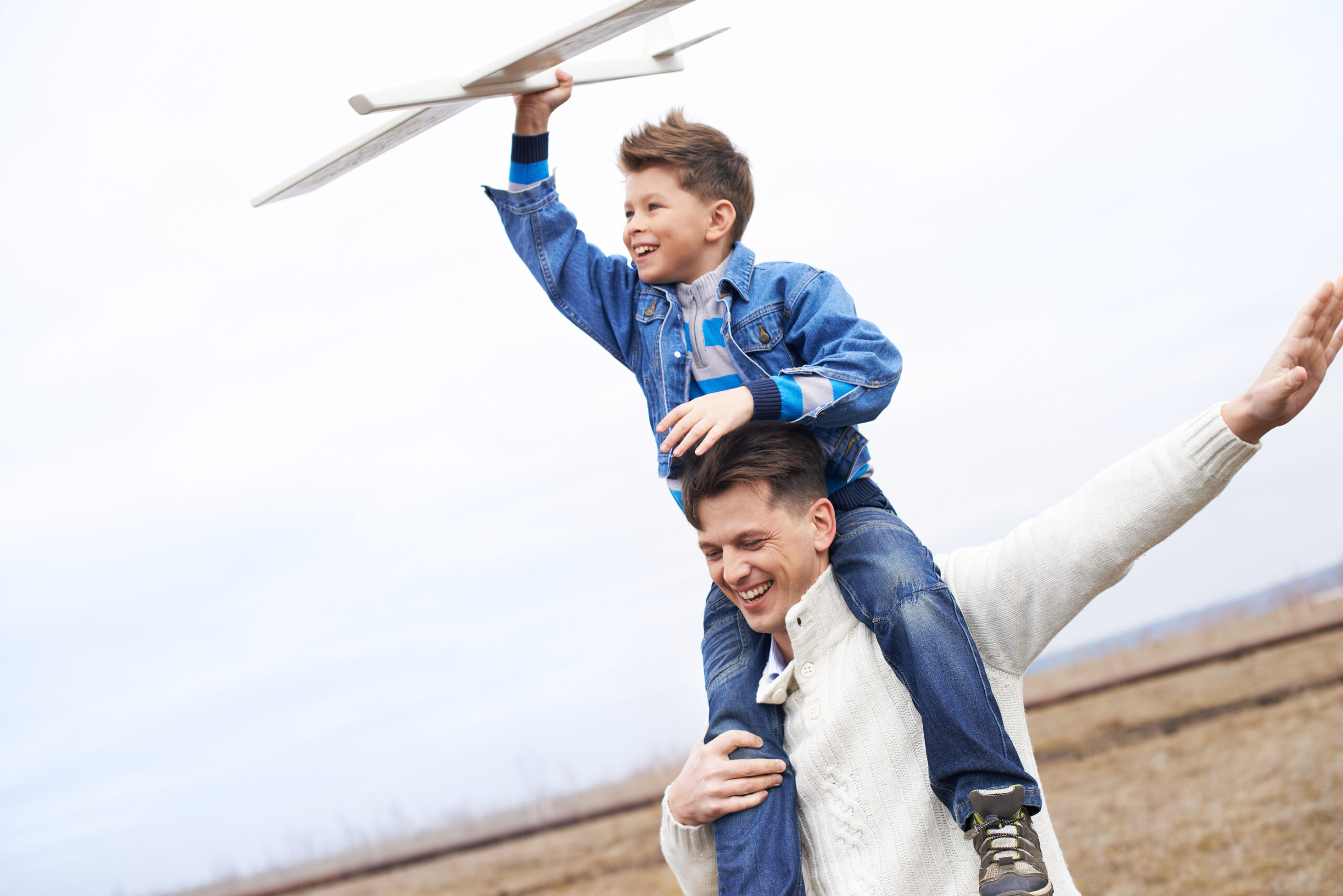 A smiling man carries a happy boy on his shoulders outdoors. The boy holds a toy airplane above his head, both wearing casual clothes. The background is a cloudy sky and an open field.