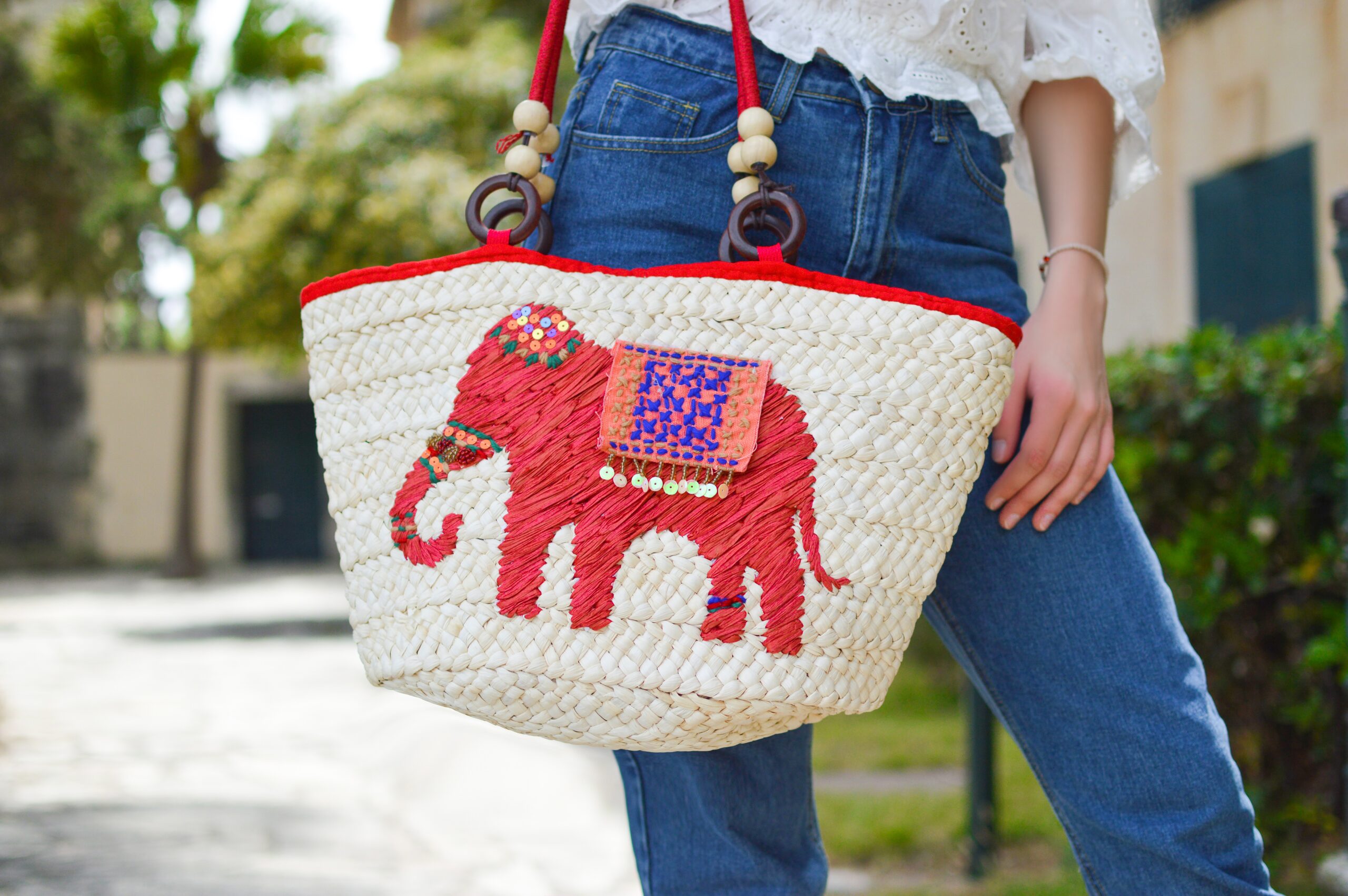 A person wearing blue jeans holds a woven straw handbag decorated with a red elephant design, colorful beads, and red trim. The background is outdoors with blurred greenery and buildings.