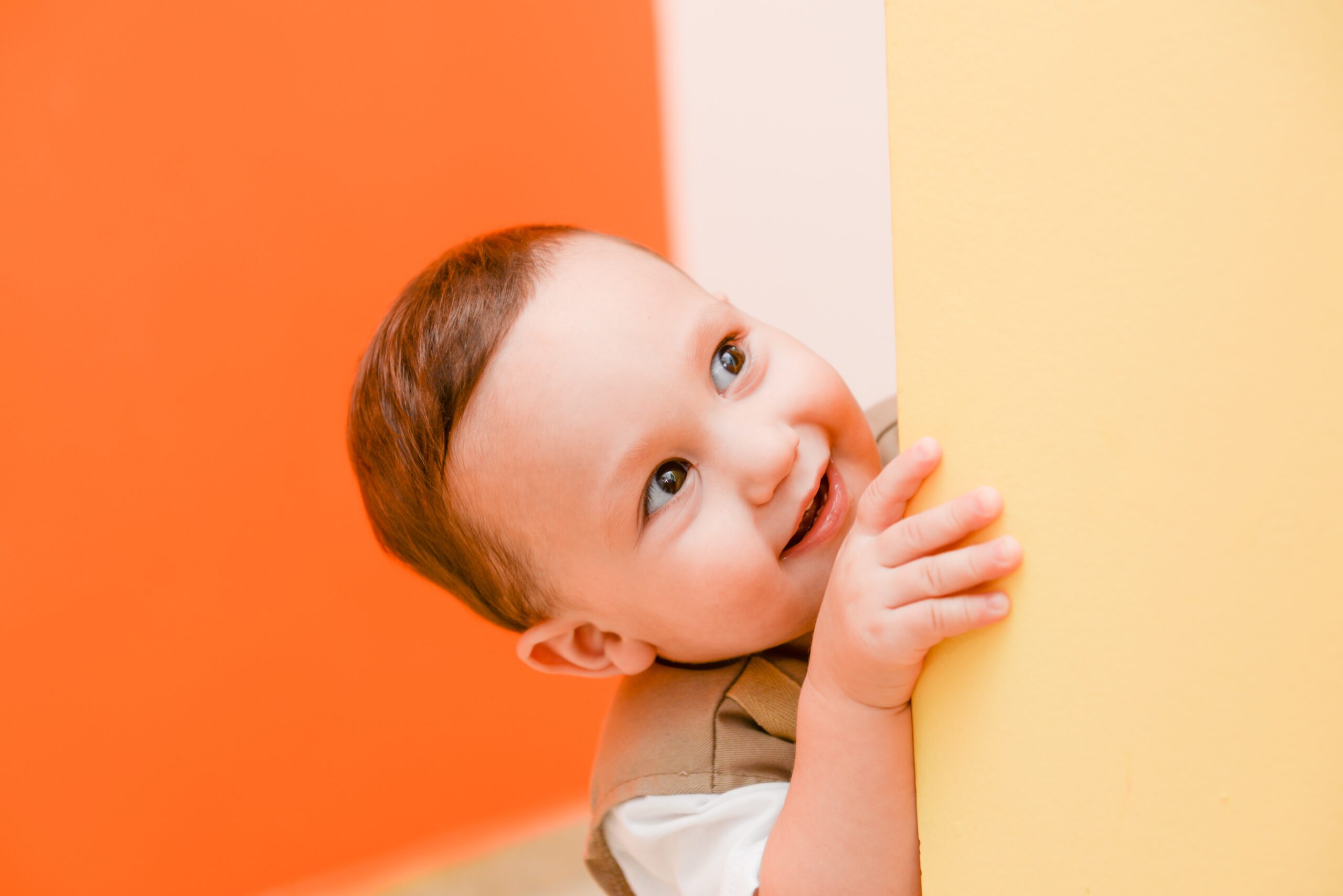 A smiling toddler with short brown hair peeks playfully from behind a yellow wall, with an orange and white background.
