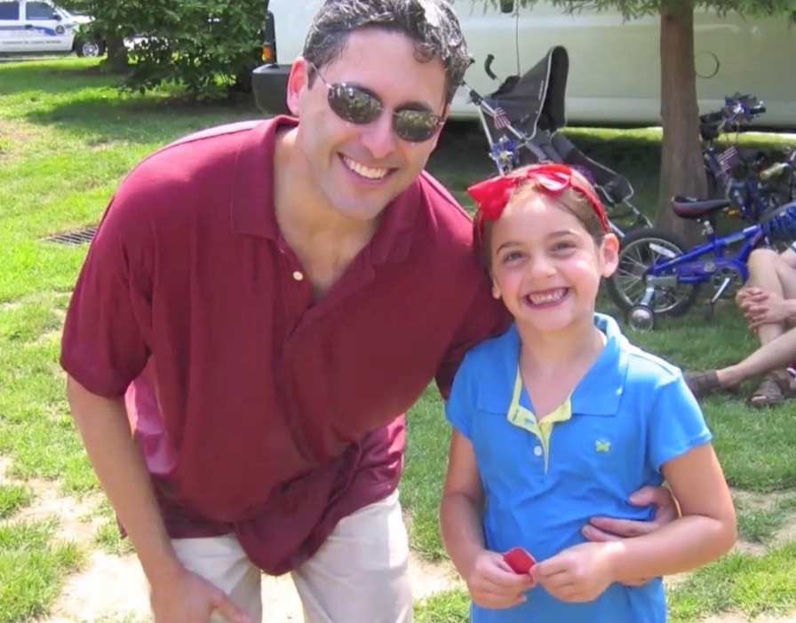 A man in sunglasses and a red shirt smiles while hugging a young girl wearing a blue shirt and red headband. Outdoors on grass with bicycles and a van nearby, they enjoy time together after ADHD Evaluations and Coaching.