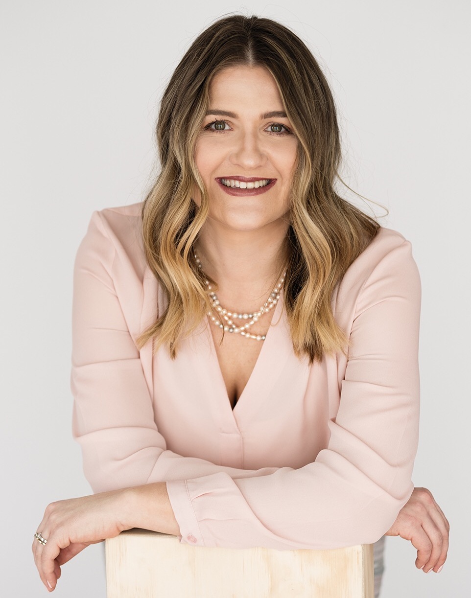 A smiling woman with wavy, shoulder-length hair wearing a light pink blouse and layered pearl necklaces leans forward with her arms resting on a light wooden surface against a plain white background.