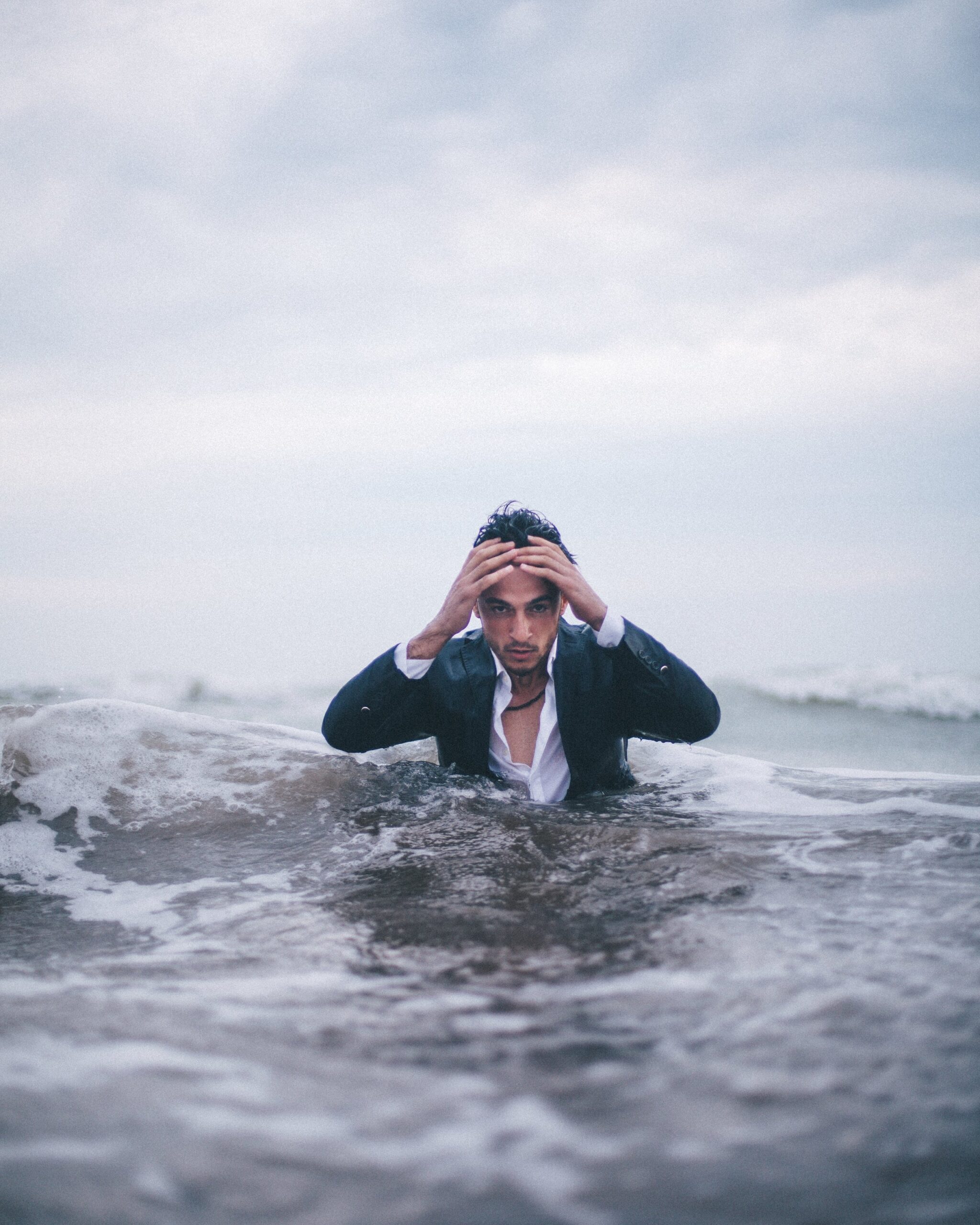 A man in a suit stands waist-deep in the ocean, holding his head with both hands, looking intently at the camera as waves crash around him under a cloudy sky.