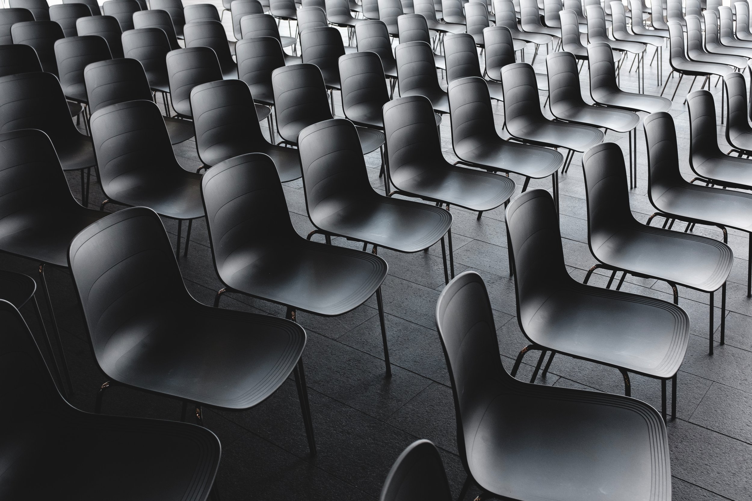 Rows of empty black plastic chairs arranged neatly in a grid pattern on a tiled floor, creating a sense of repetition and symmetry. The chairs are unoccupied, suggesting a pause before an event.