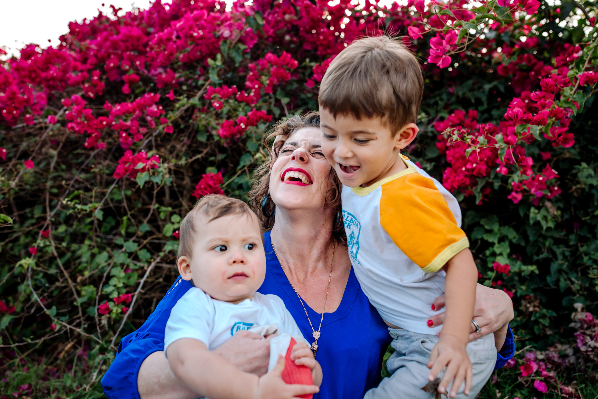 A smiling woman in a blue shirt holds two young children in front of vibrant pink flowers. One child smiles and hugs her neck while the other sits on her lap with a curious expression.