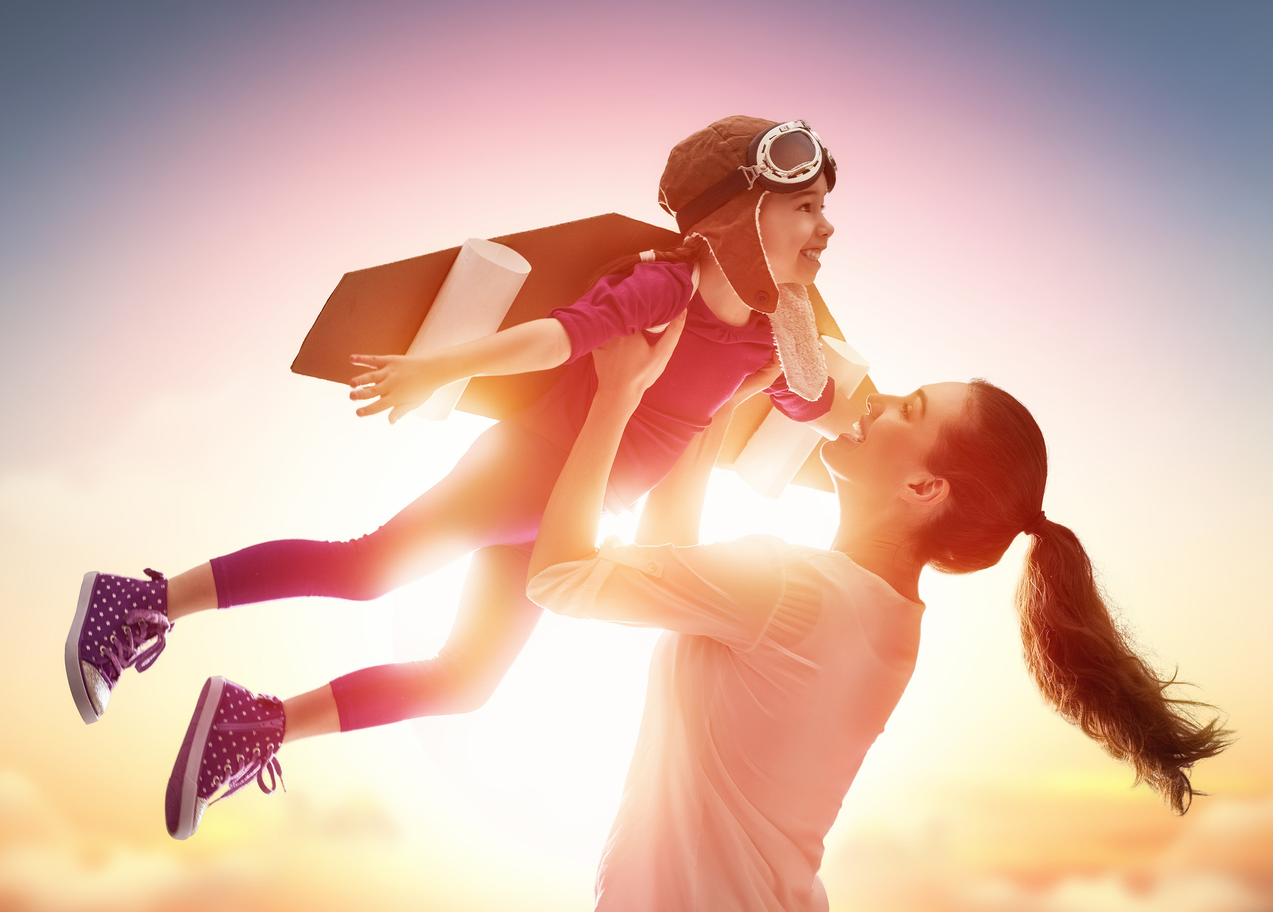 A woman lifts a smiling child into the air at sunset. The child wears a toy jetpack, aviator hat, and goggles, pretending to fly while the woman smiles up at them. Both are backlit by a warm, glowing sky.