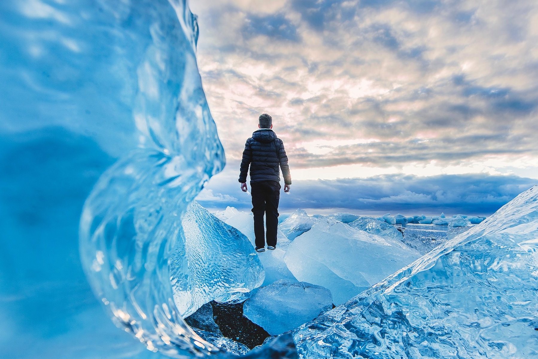 A person in a dark jacket stands on large blue ice chunks, surrounded by jagged icy formations, under a dramatic cloudy sky at sunset or sunrise.