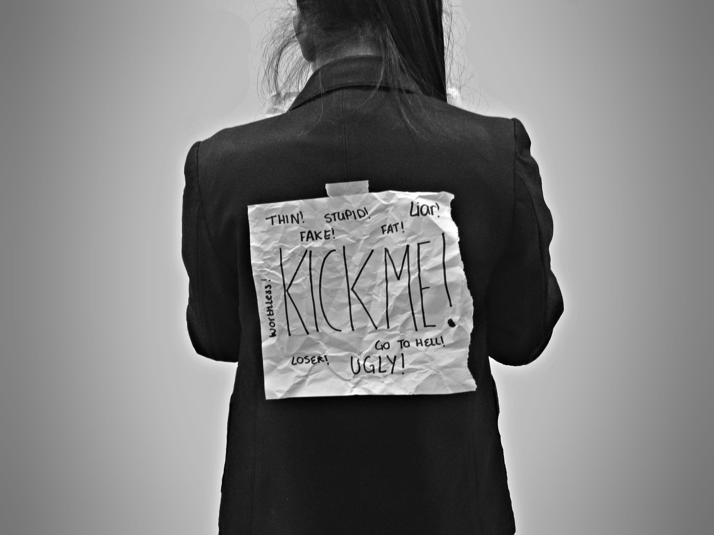 A person with long hair, seen from behind, wears a suit jacket with a crumpled paper taped to their back. The paper reads KICK ME! and is surrounded by insults like stupid, loser, fat, and go to hell.