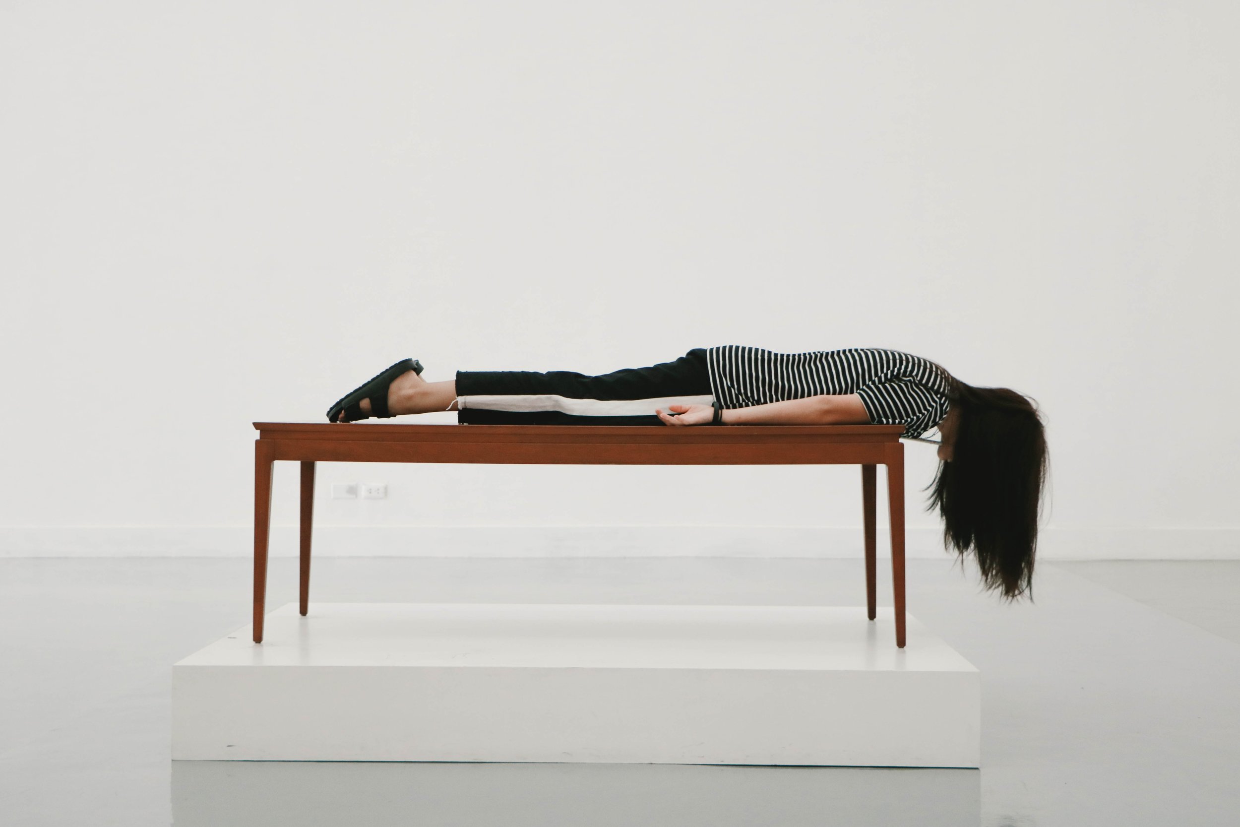 A person wearing black pants and a striped shirt lies face down and straight on a wooden table, with long hair hanging off the edge, in a minimalist white room.