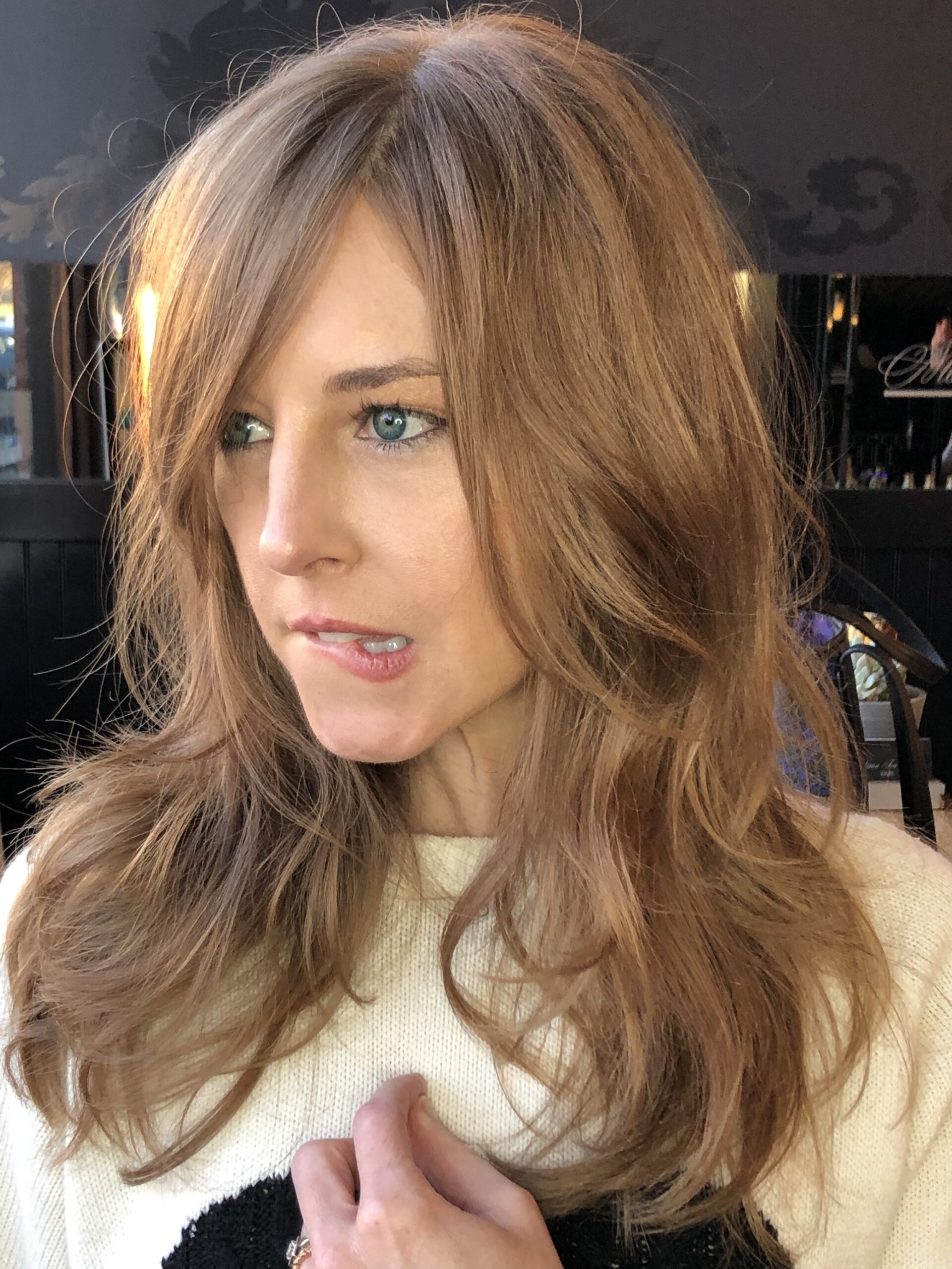 A woman with long, wavy, light brown hair and blue eyes wears a cream sweater and looks slightly to the side in soft natural light, with a faint smile on her lips.