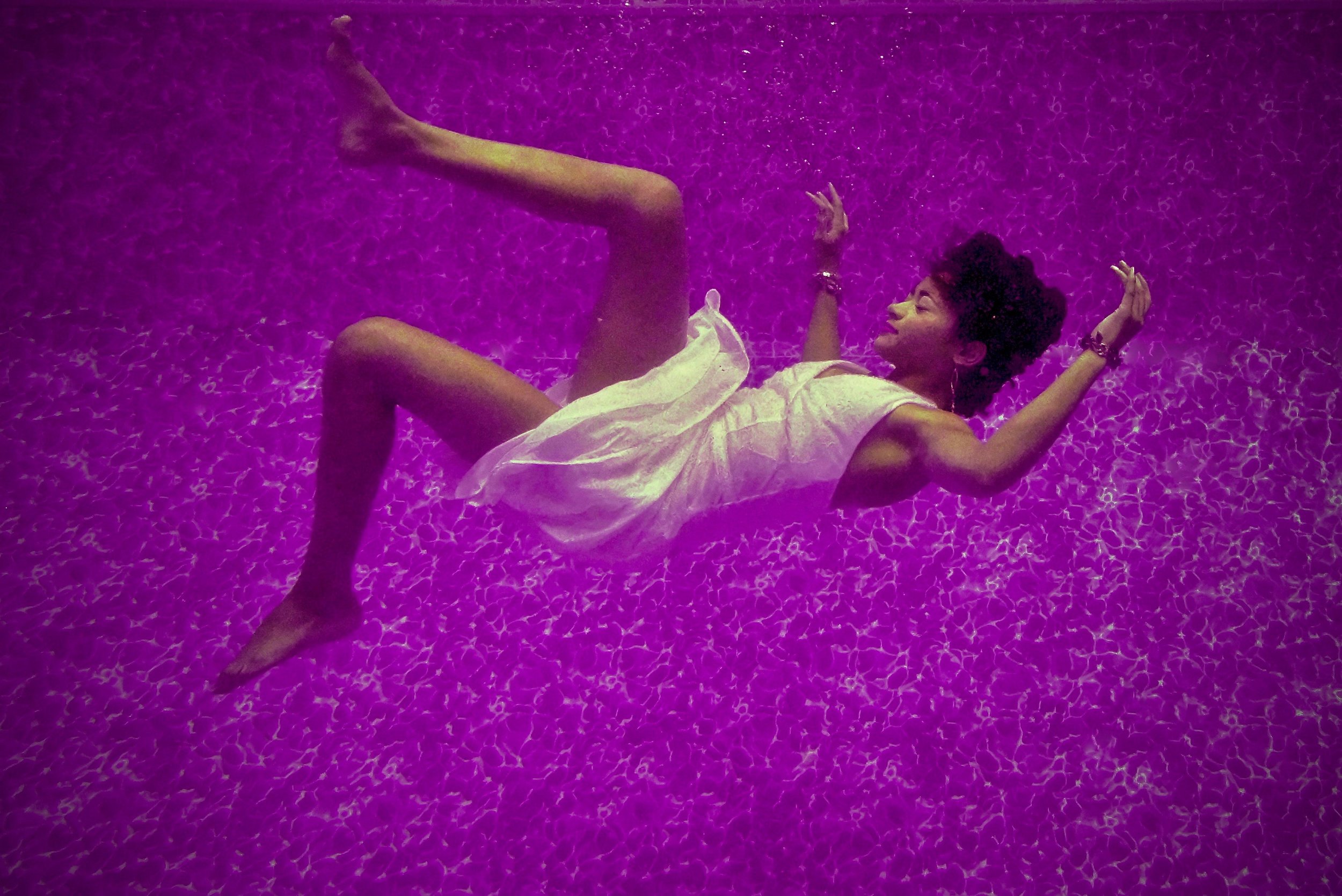 A woman in a white dress floats underwater in a swimming pool with a purple tint, arms and legs bent, looking upwards, surrounded by shimmering patterns on the pool floor.