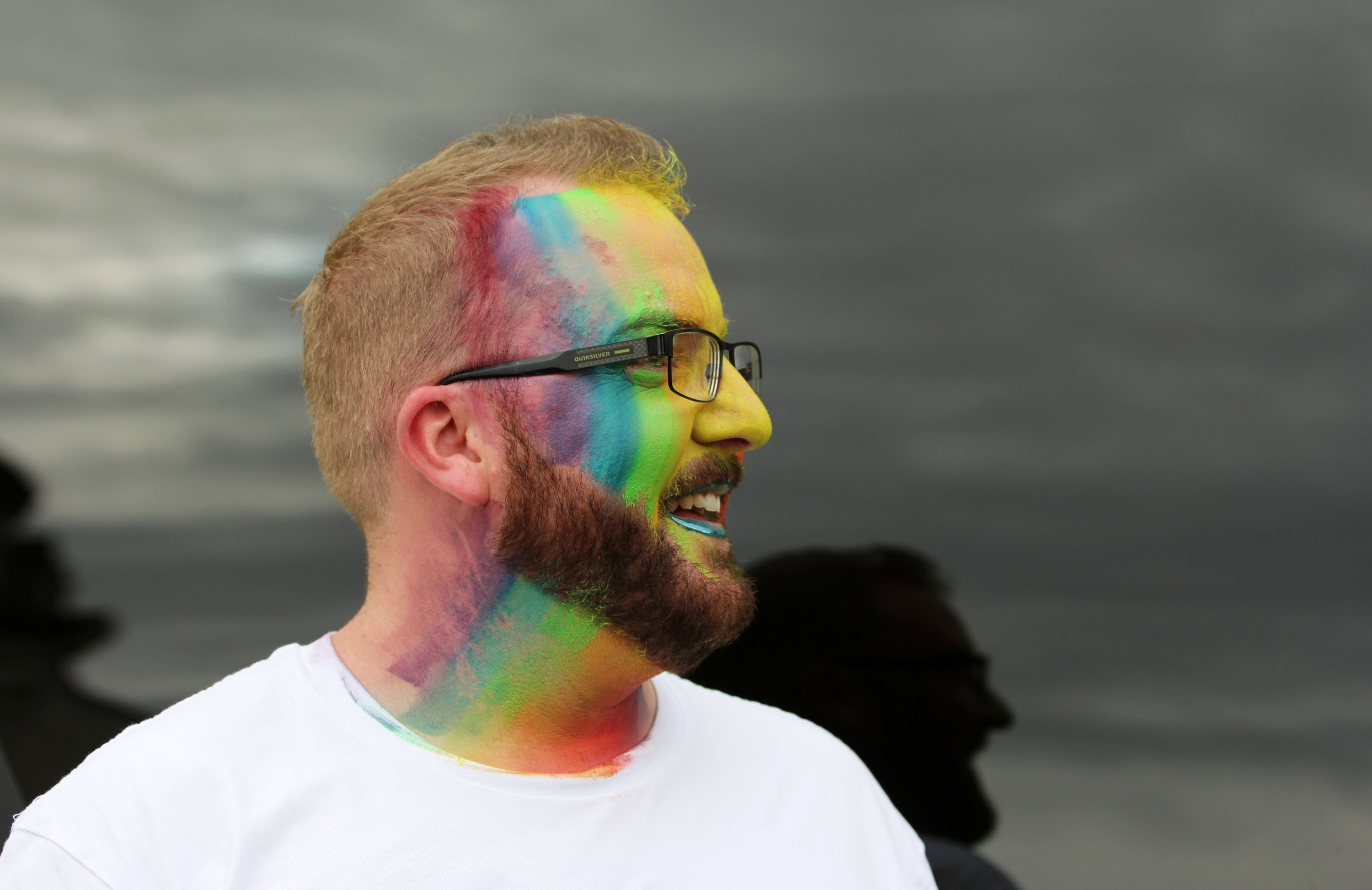 A bearded person with glasses and short blond hair smiles, with their face and neck painted in rainbow colors, standing against a dark, cloudy background.