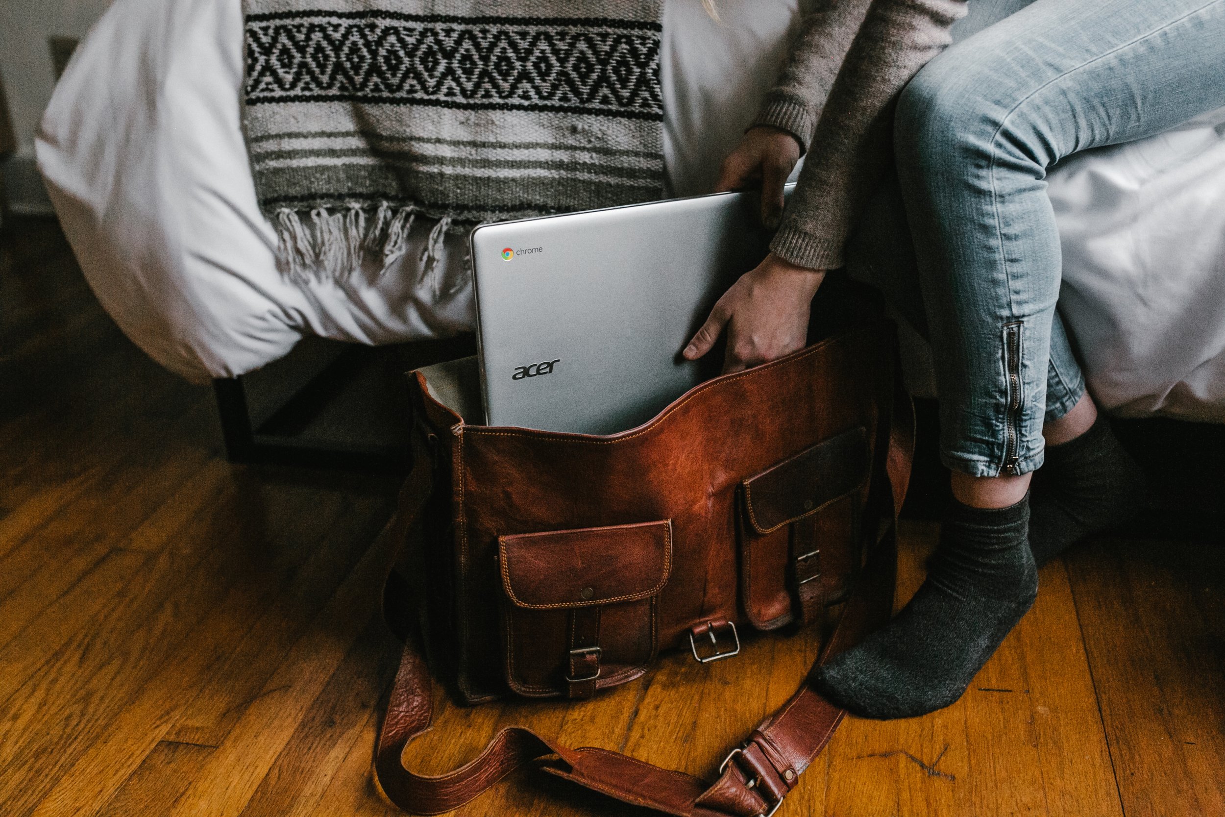 A person sits on the edge of a bed, placing a silver Acer Chromebook into a brown leather satchel on a wooden floor. Only their hands and legs are visible.