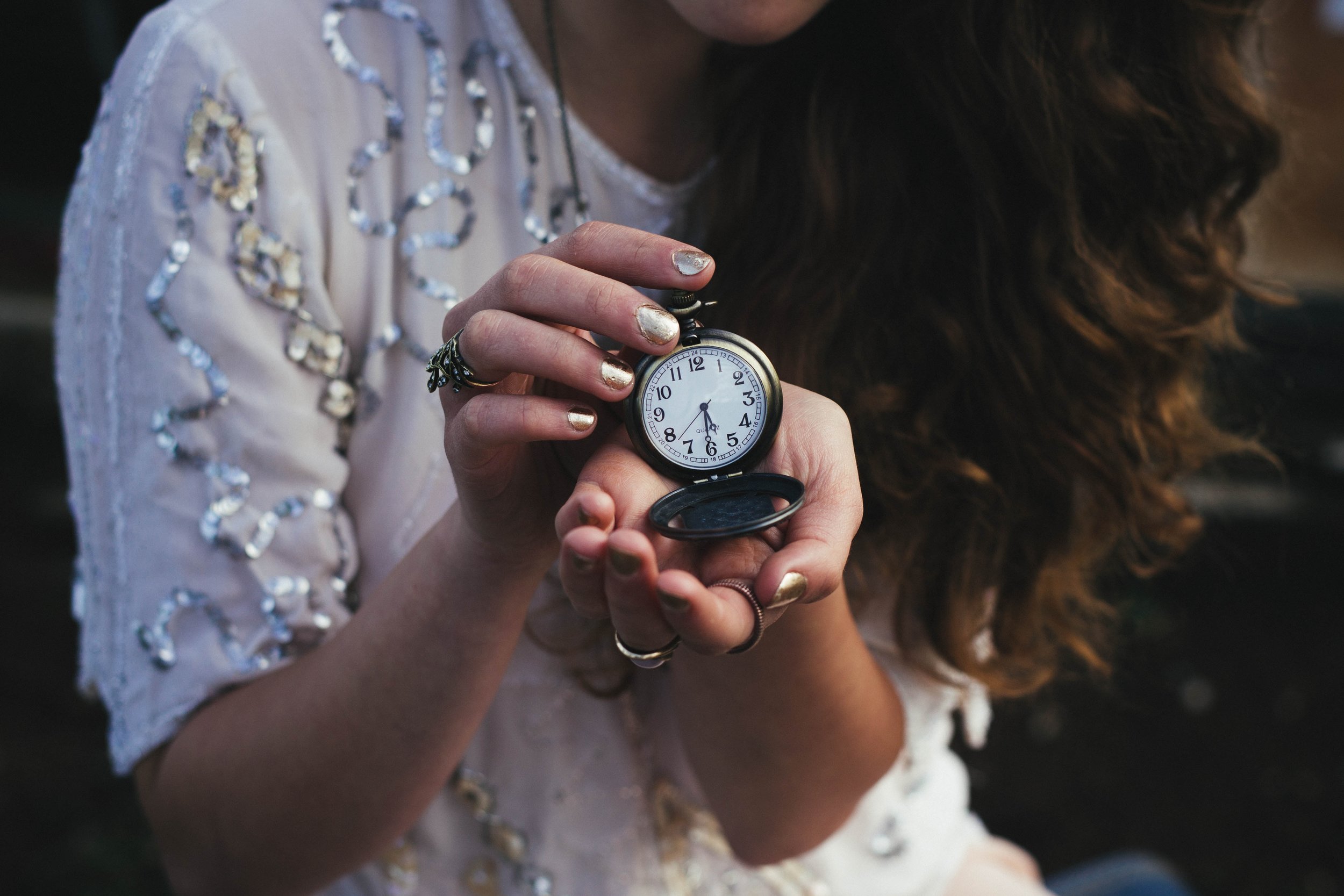 A person with wavy brown hair, wearing a white beaded top, holds an open pocket watch in their hands. The watch shows the time as 4:34. Their nails are painted metallic silver.