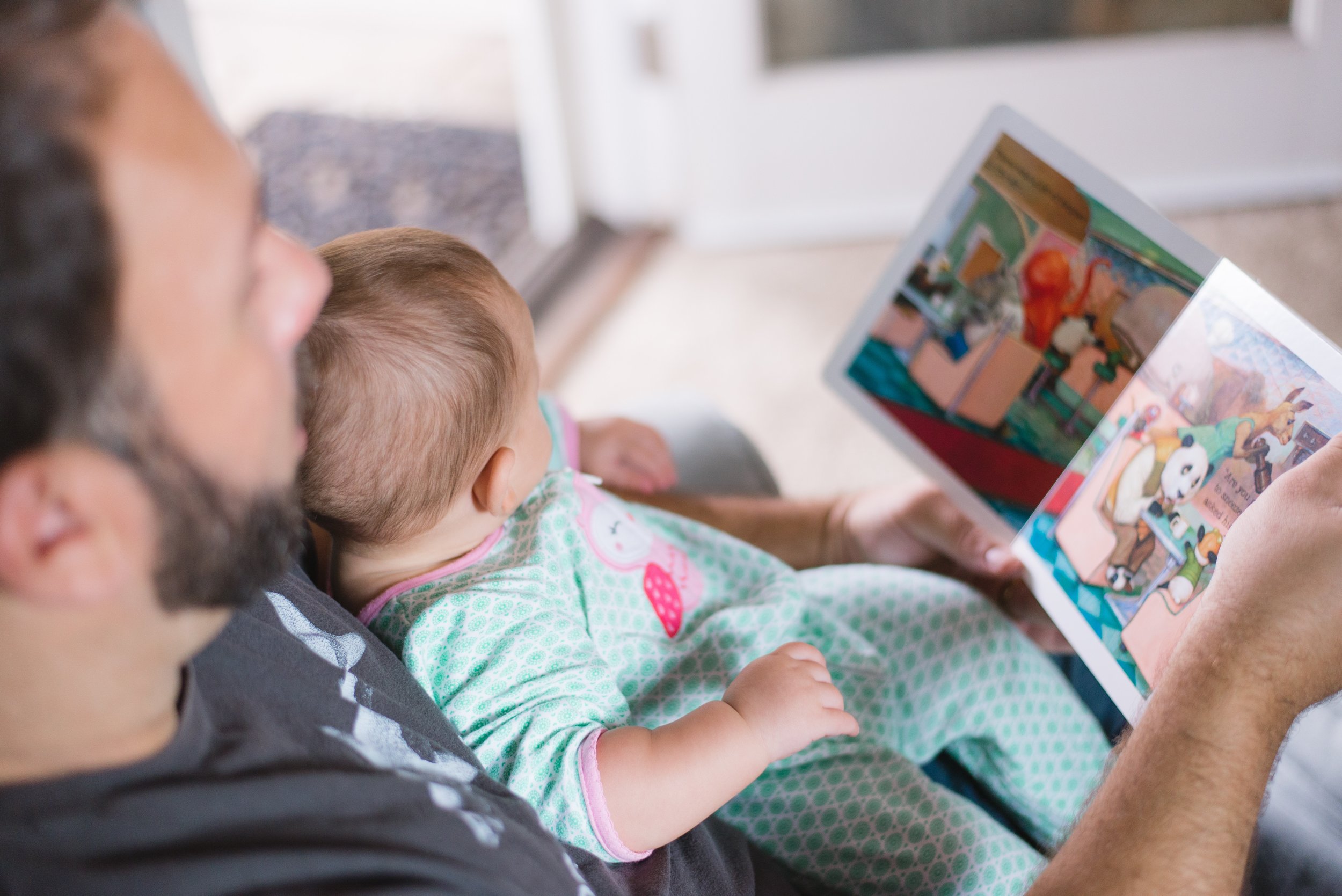 A man and a baby sit close together as the man reads a colorful picture book to the baby, who is wearing green patterned pajamas. The scene appears cozy and intimate, with natural light in the background.