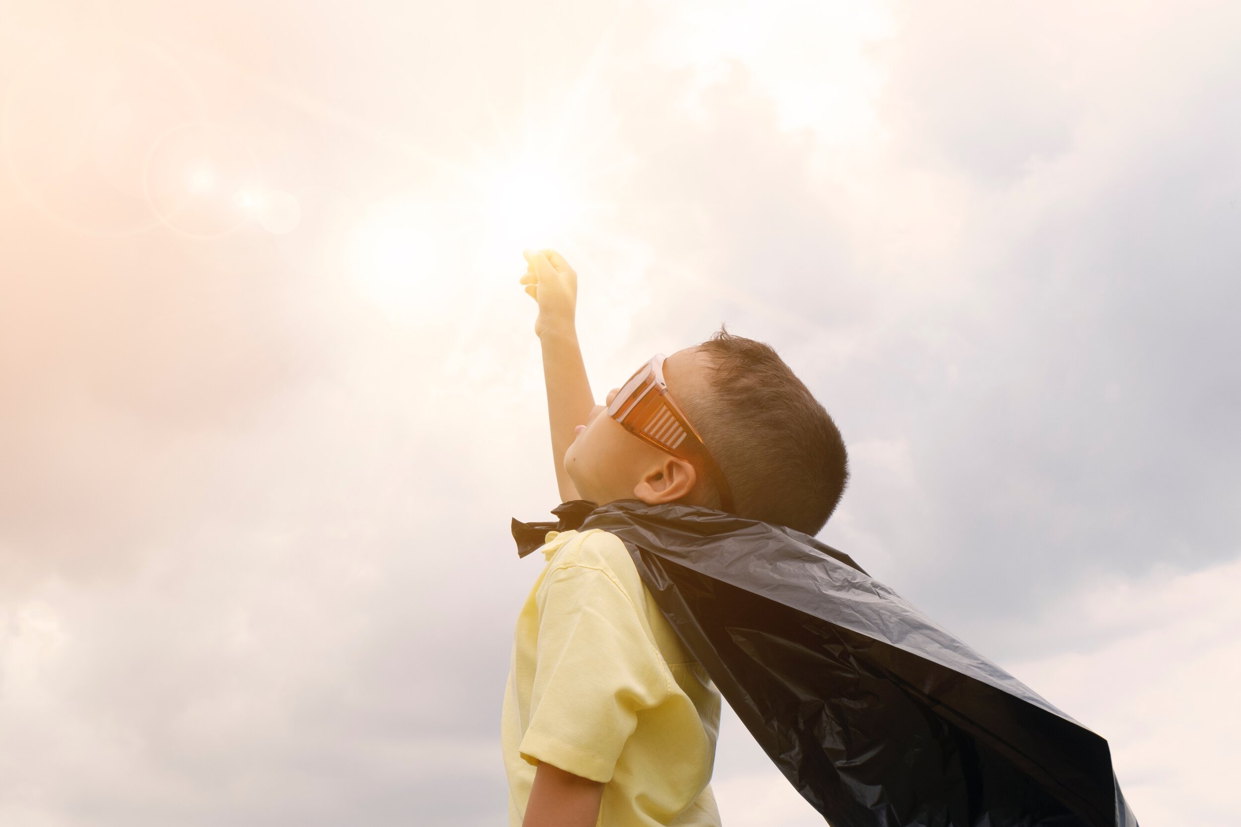 A young boy wearing a black cape and sunglasses raises his fist toward the bright sun, posing as if he is a superhero, with a cloudy sky in the background.