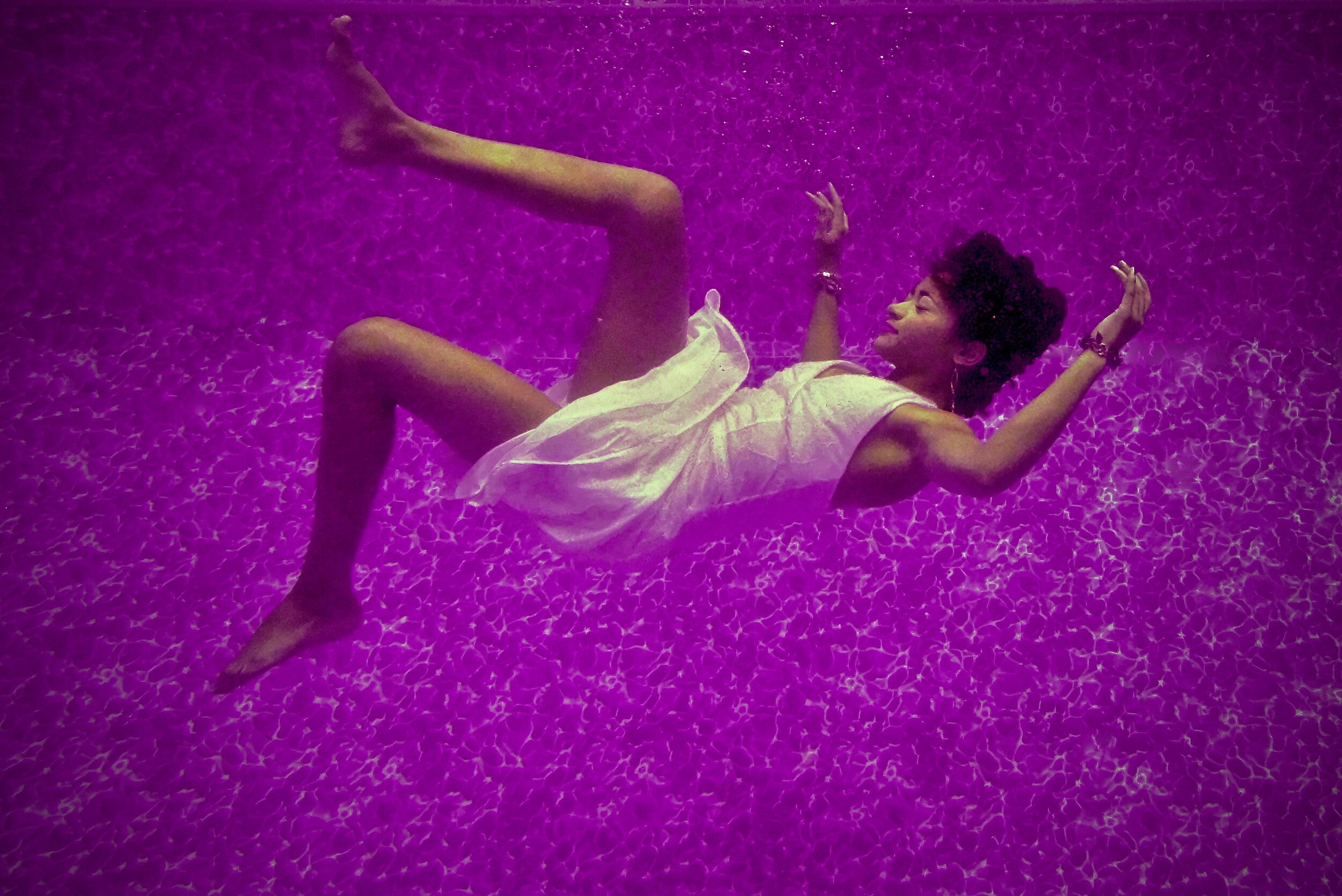 A person in a white dress floats underwater in a purple-lit pool, arms and legs extended in different directions, appearing weightless and serene.