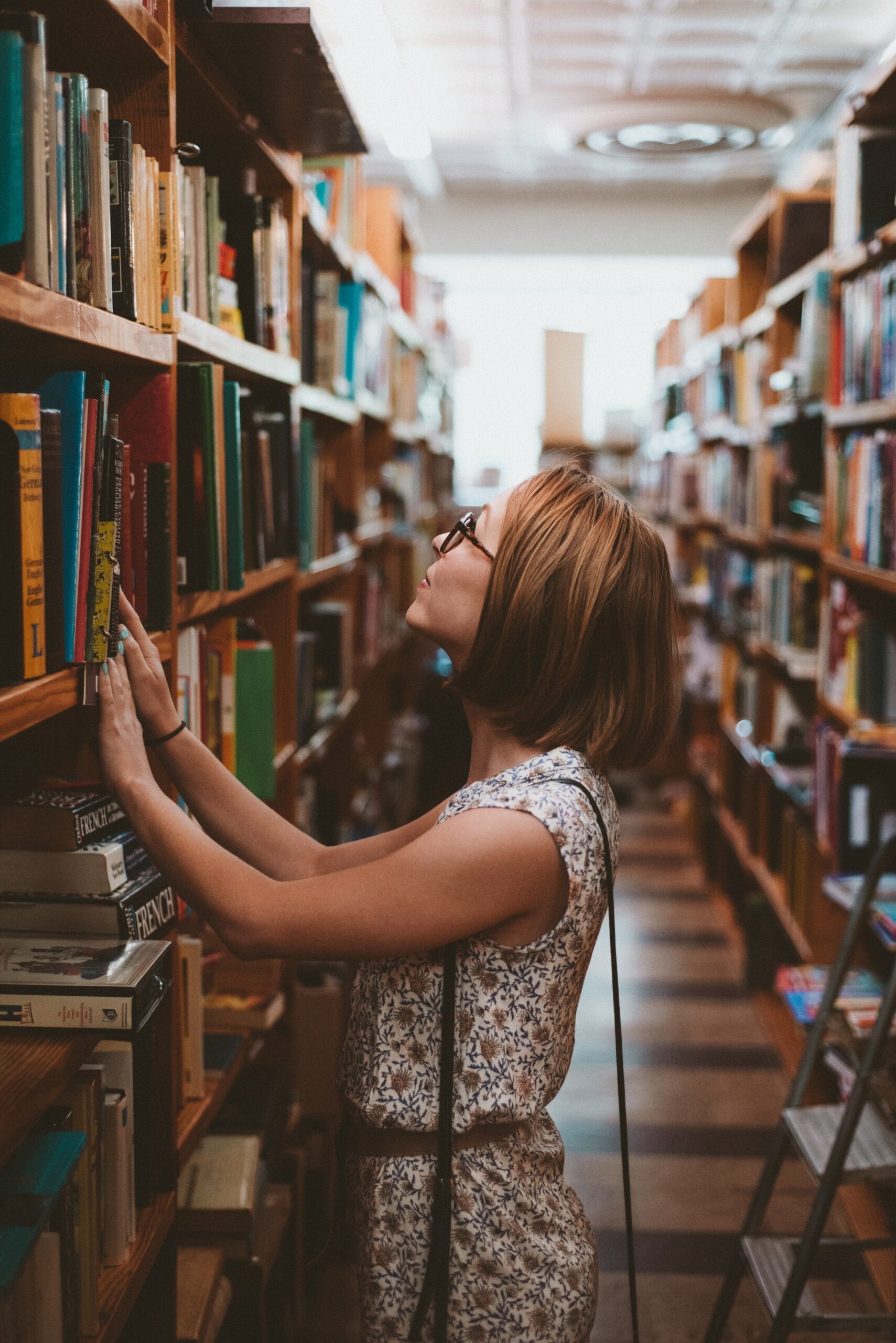 A woman wearing glasses and a sleeveless floral outfit browses books on a tall shelf in a library or bookstore, surrounded by rows of bookshelves.