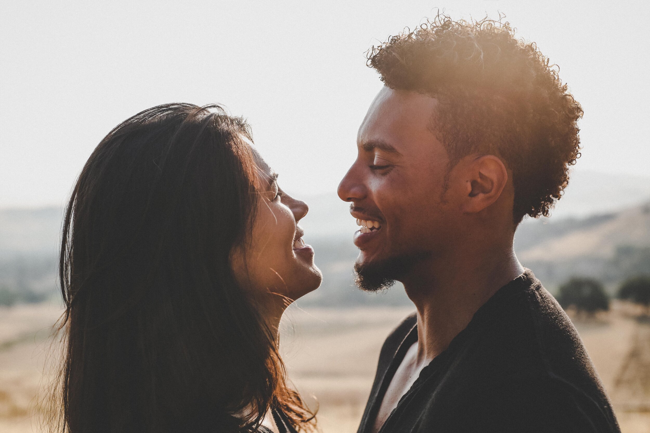 A woman and a man stand close together outdoors, smiling and looking into each others eyes, with sunlight shining behind them and blurred nature in the background.