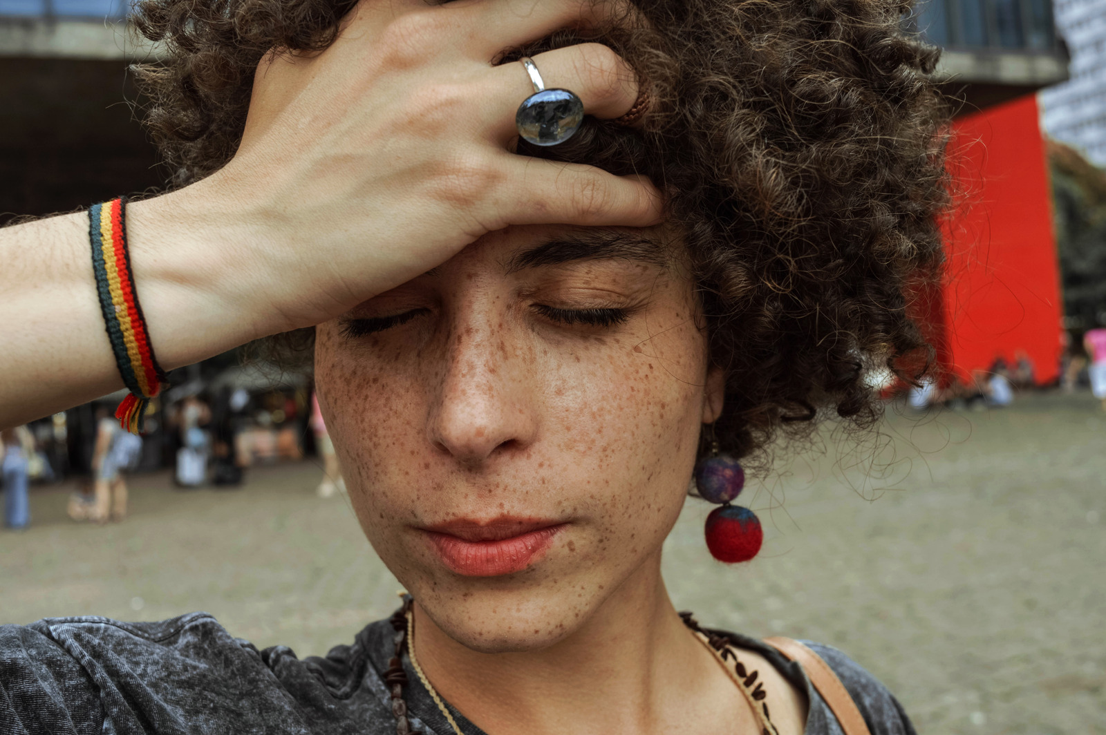A person with curly hair and freckles touches their forehead with closed eyes, wearing a ring, colorful earring, and a woven bracelet, standing outdoors in an urban area.