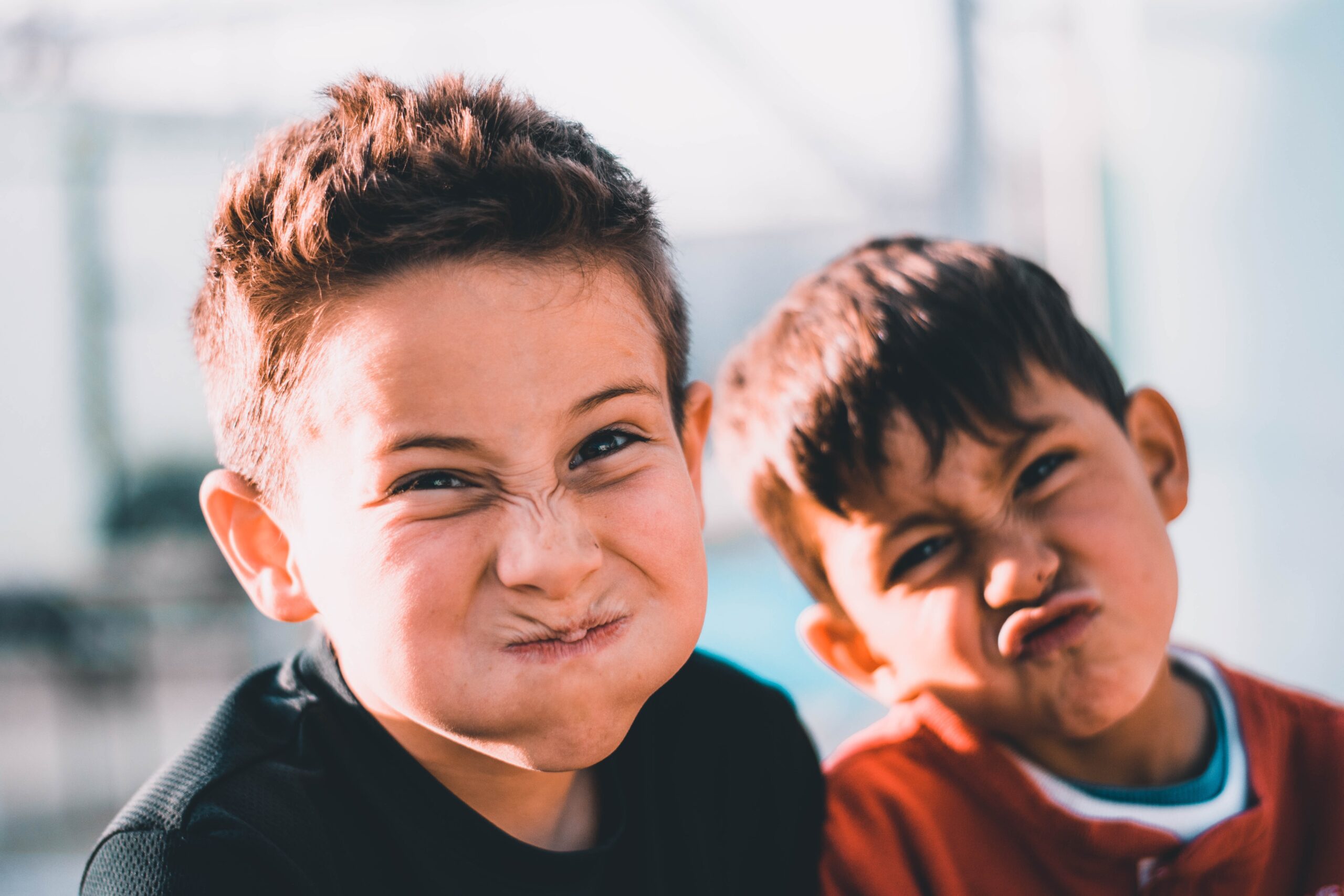 Two young boys are making funny faces at the camera, puffing their cheeks and scrunching their noses. They are close together, smiling playfully, with a blurred outdoor background behind them.