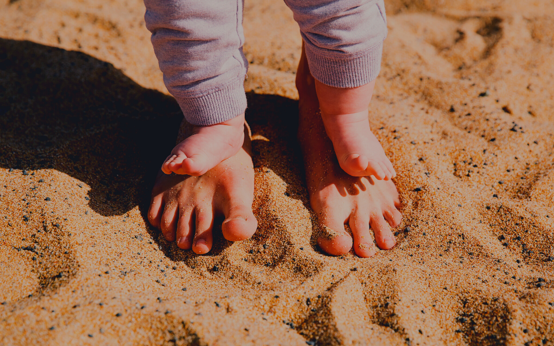 A baby standing on an adults bare feet in the sand, both pairs of feet close together. The adults feet are partially covered by the babys feet, and both are surrounded by sunlit sand.
