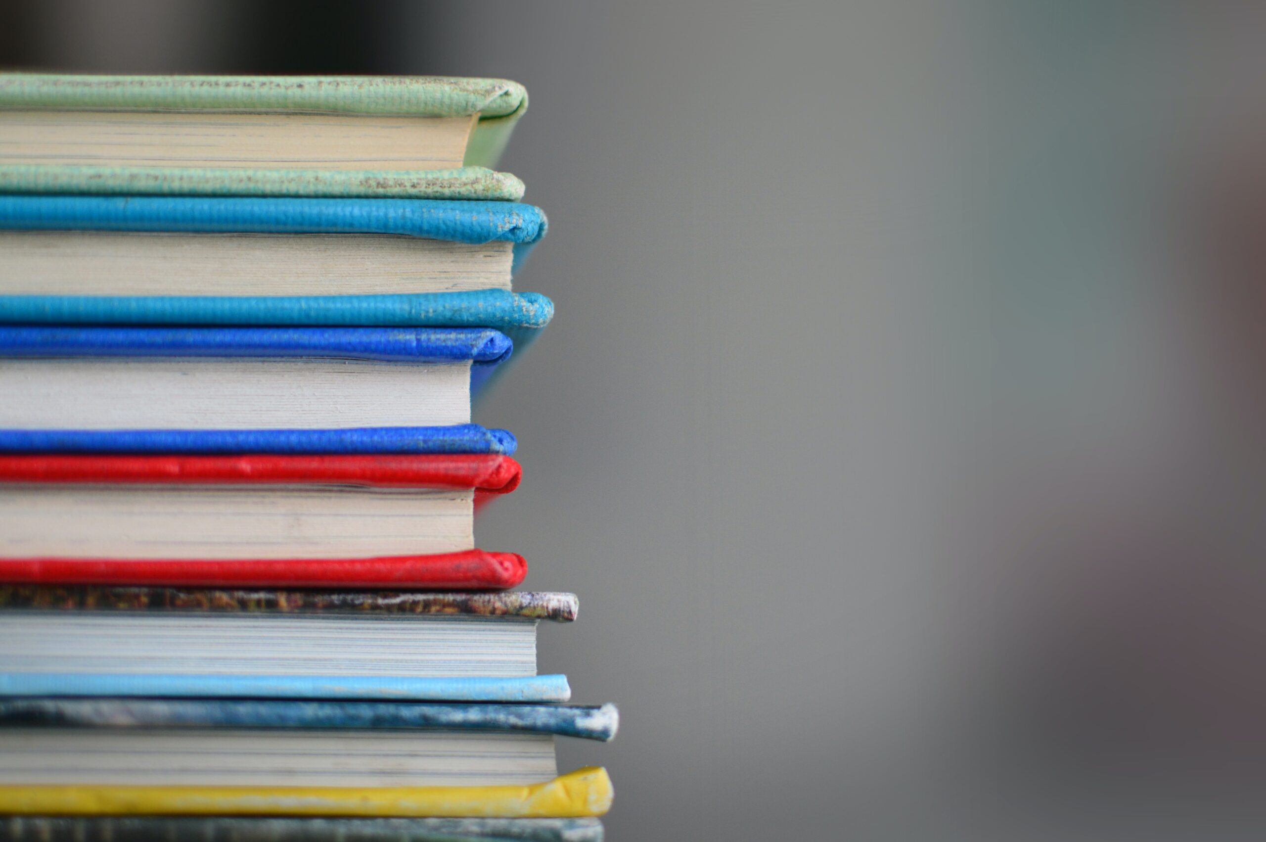 A close-up of a stack of books with colorful covers and spines, including blue, red, yellow, and green, arranged on the left side of the image against a blurred gray background.