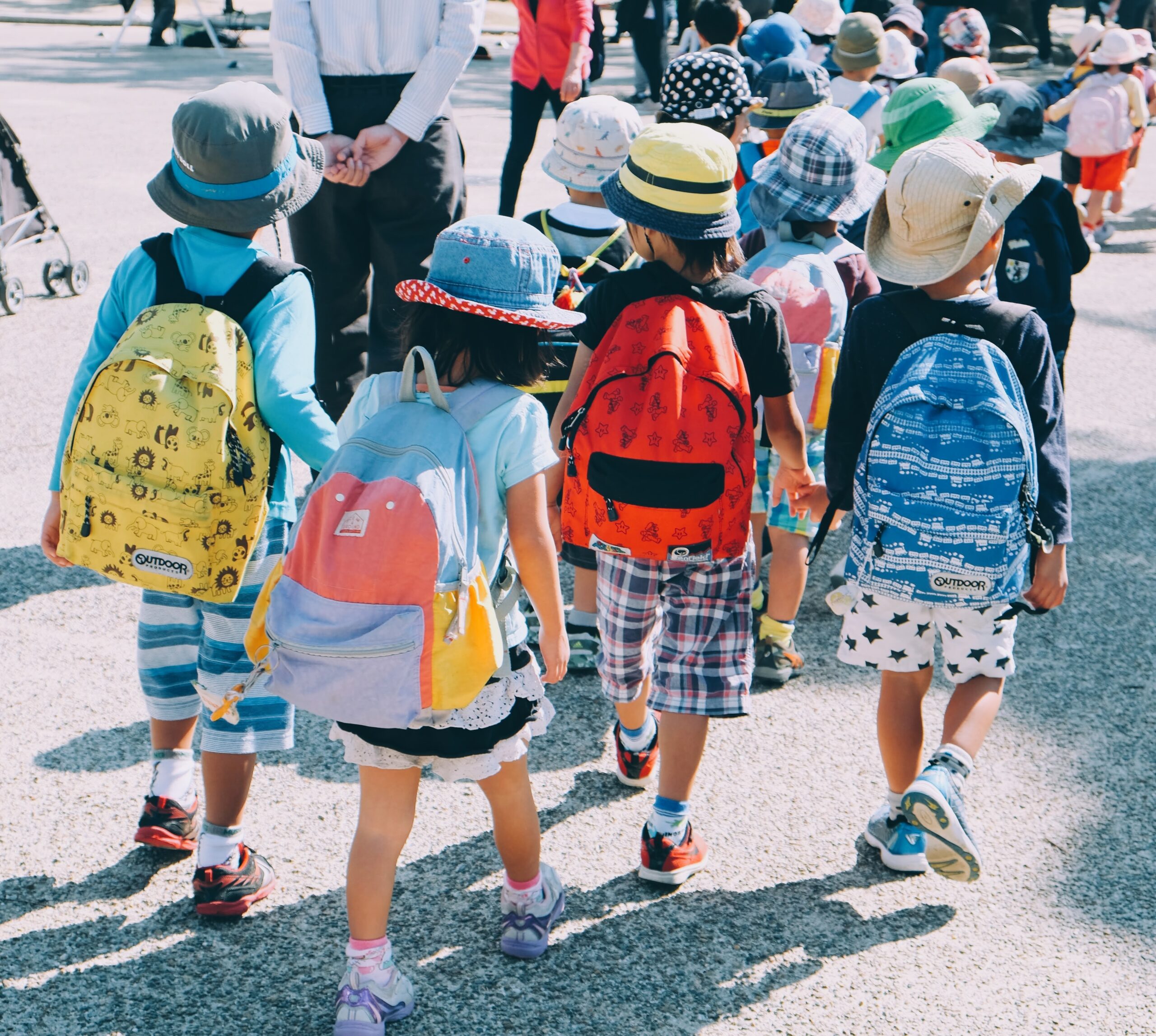 A group of young children wearing colorful hats and backpacks walk together outdoors on a sunny day, holding hands, with more children and adults visible in the background.