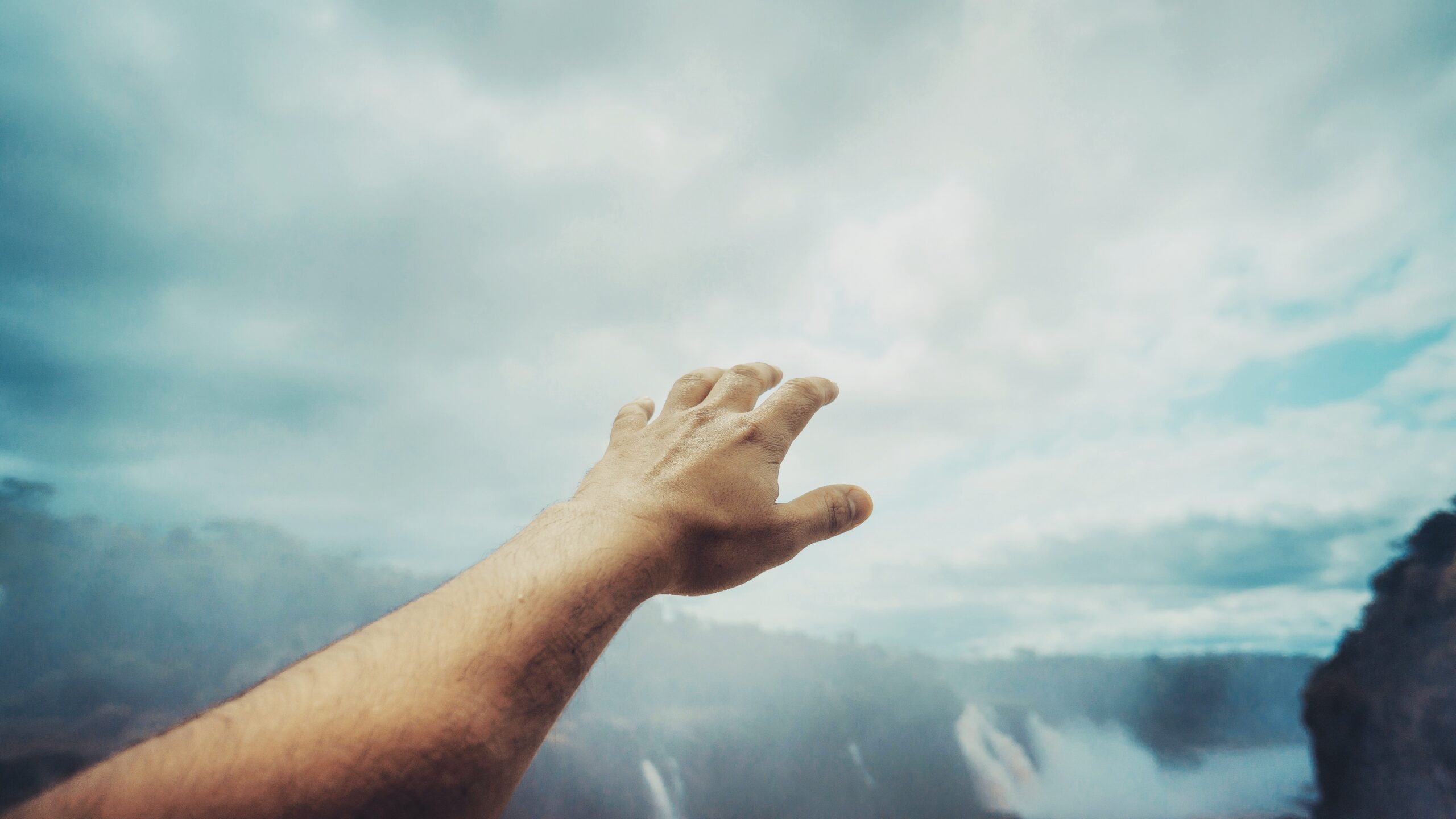 A person’s outstretched hand reaches toward a cloudy sky above a misty landscape with waterfalls and distant trees, creating a sense of longing or aspiration.