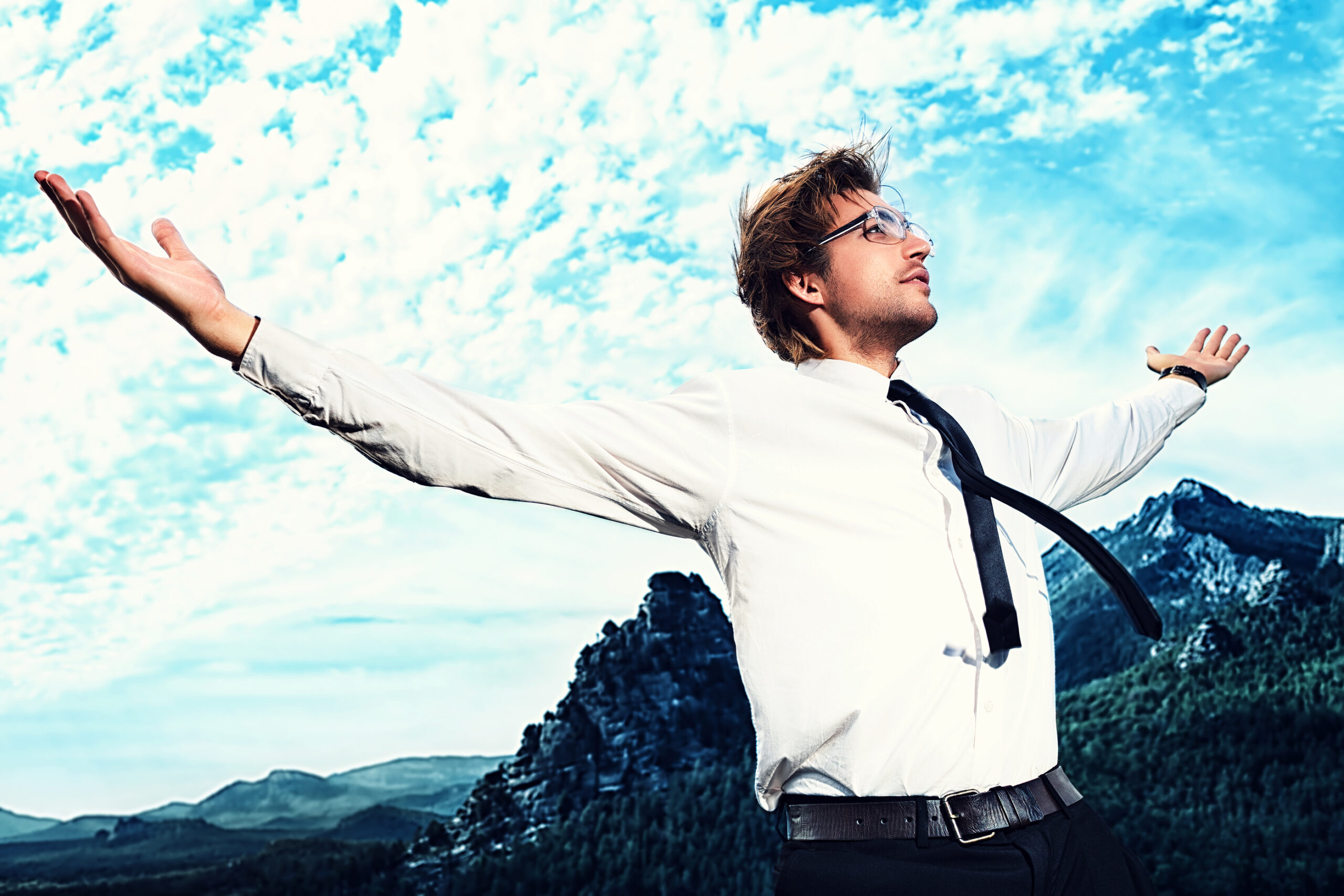 A man in a white shirt and tie stands outdoors with arms outstretched, looking upward. Mountains and a cloudy blue sky are in the background, giving a sense of freedom and achievement.