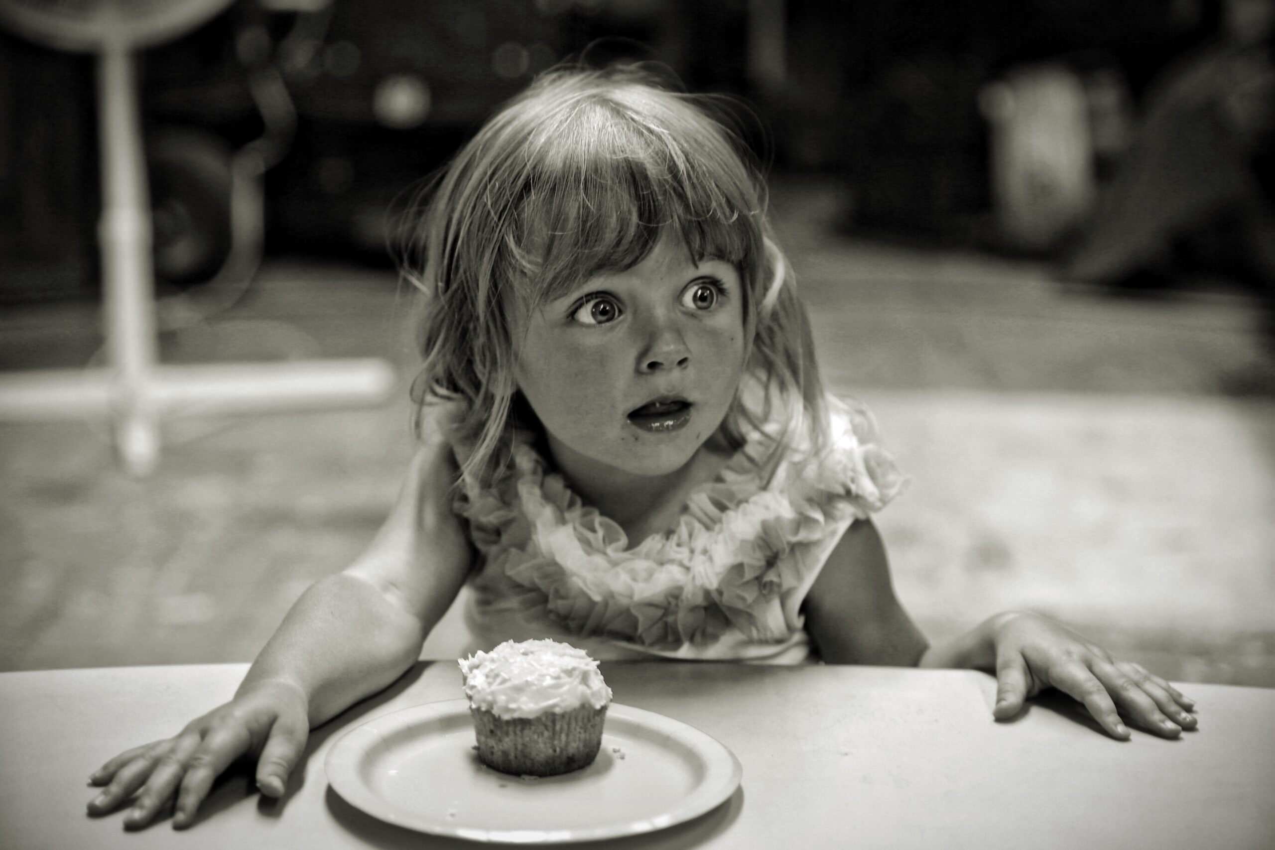 A young child with wide eyes and an amazed expression sits at a table, leaning forward with hands on the surface. There is a single frosted cupcake on a plate in front of them. The image is in black and white.