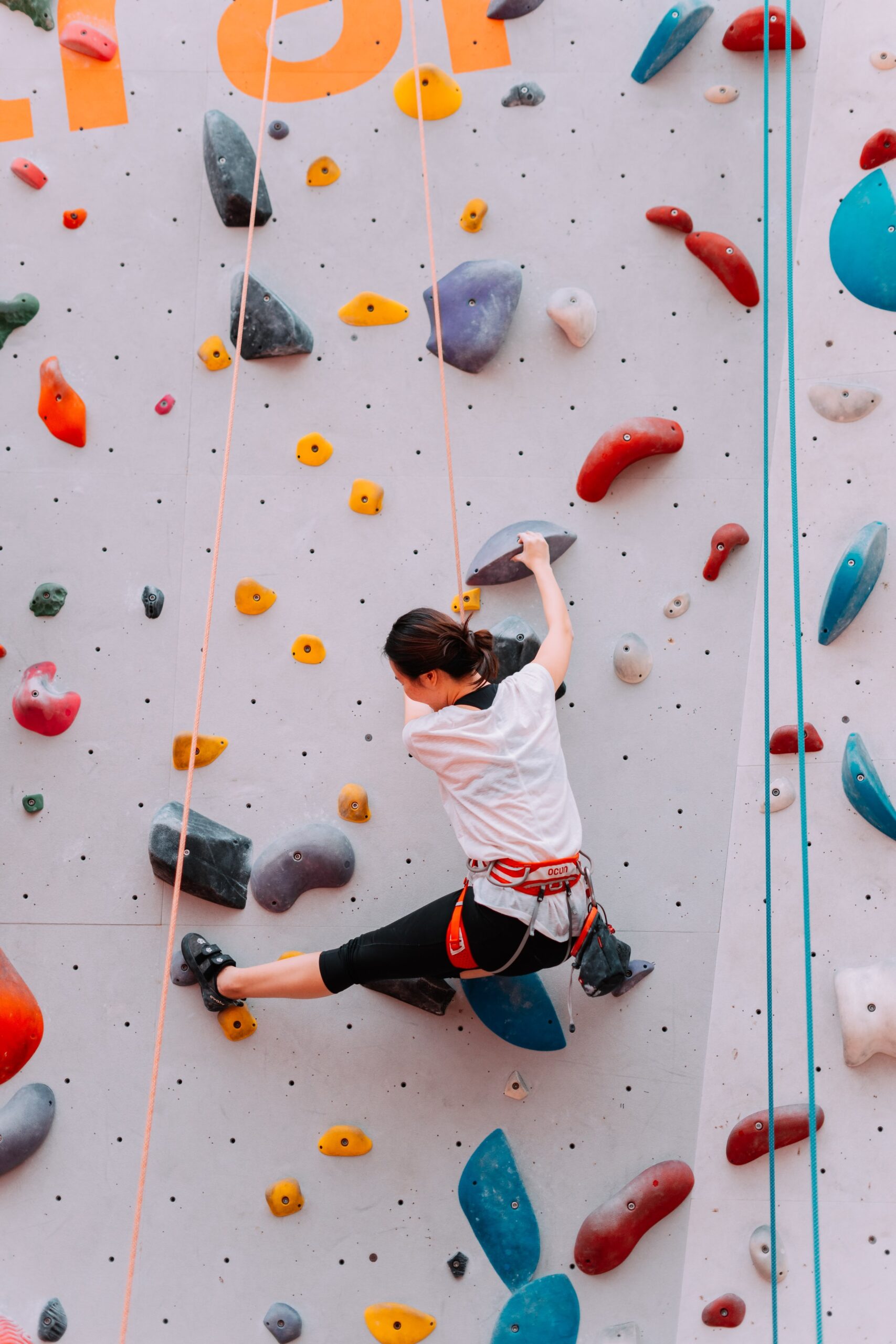 A person wearing a harness and climbing shoes scales an indoor rock climbing wall with colorful holds. They are reaching up with one hand and have one leg stretched out for support.