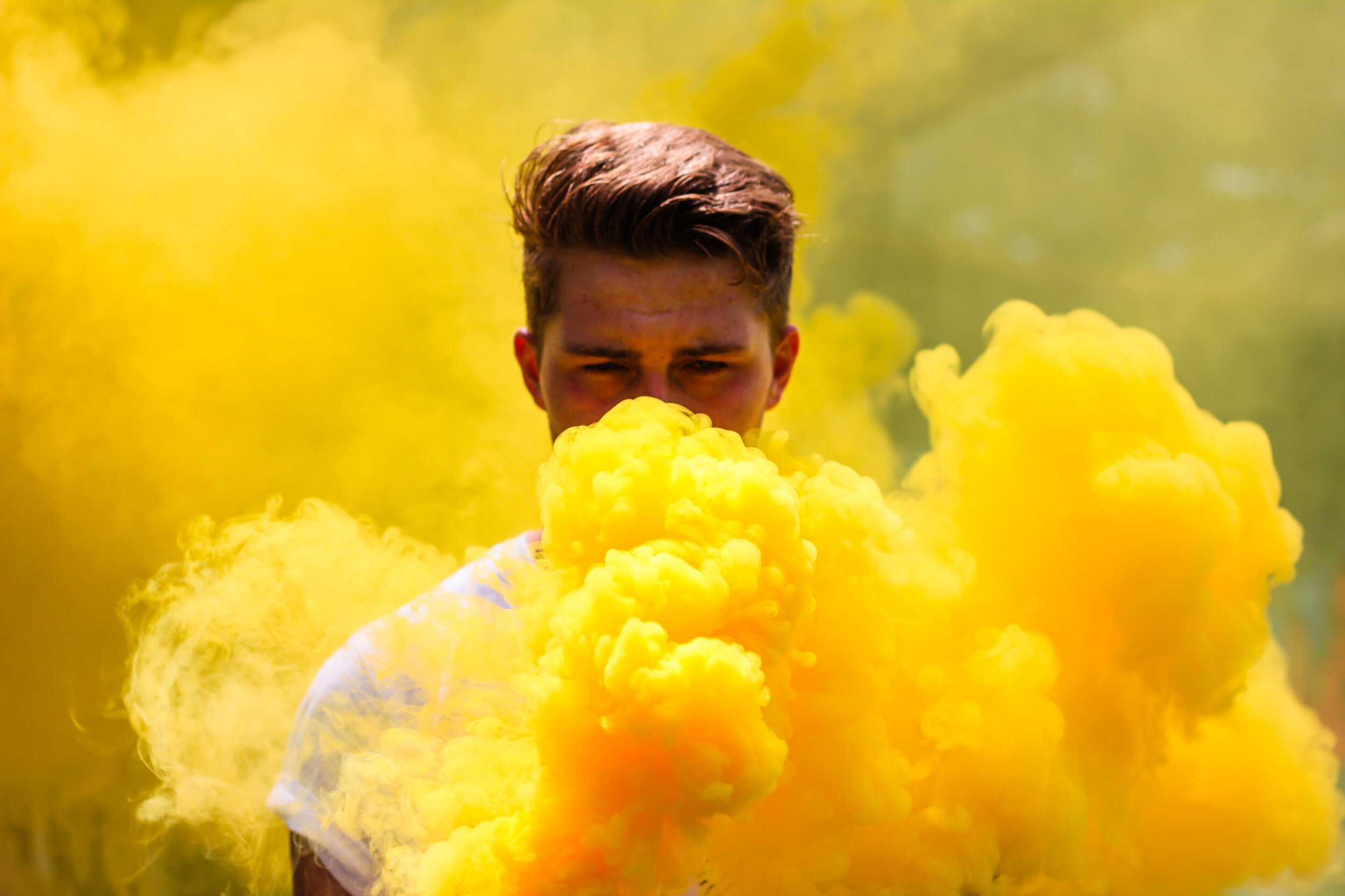A person with light brown hair is standing outdoors, partially obscured by thick yellow smoke surrounding their face and upper body. The background is blurred with a yellow haze.