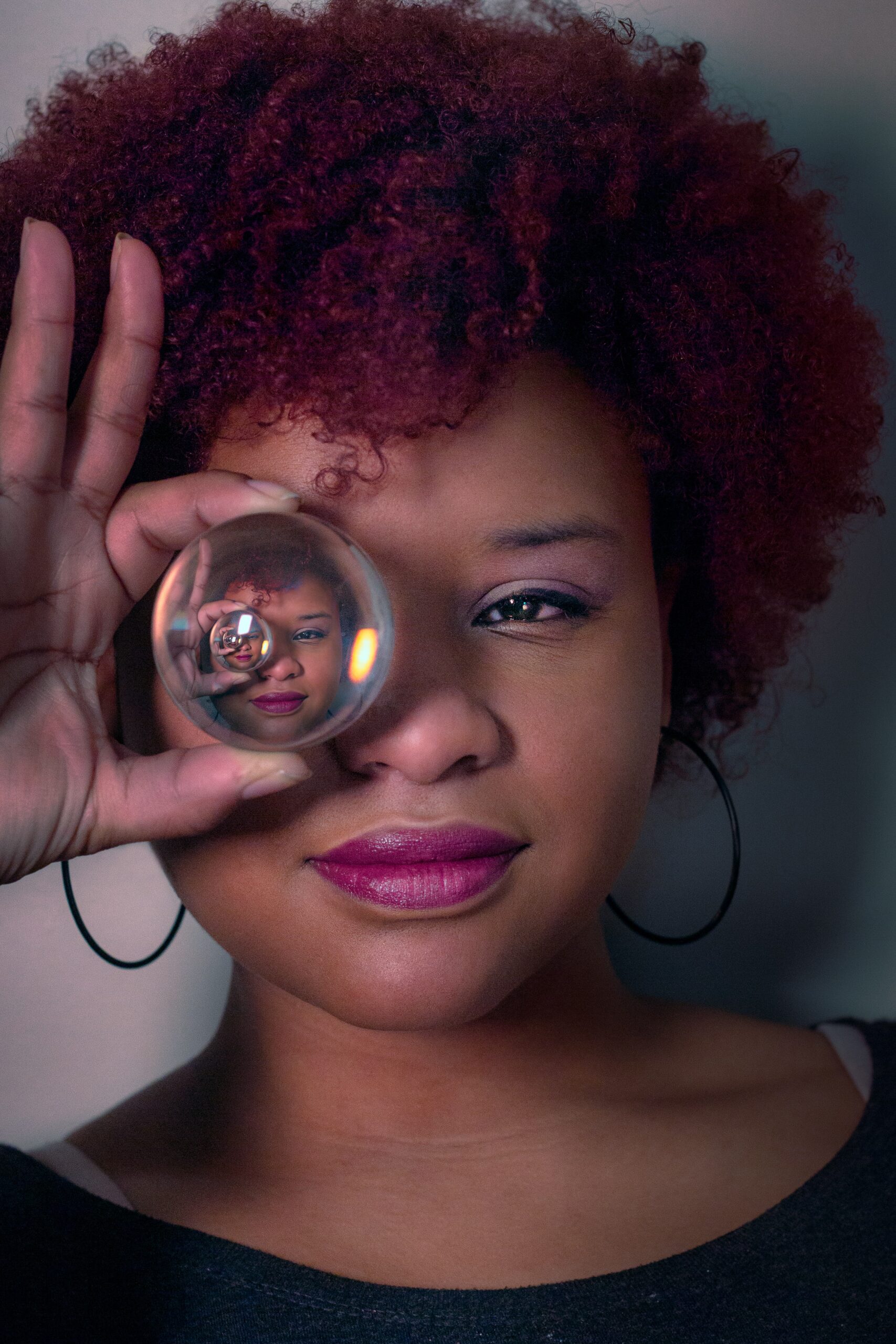 A person with curly red hair holds a glass sphere over one eye, reflecting their face inside the sphere. They wear hoop earrings and a dark top, and have a slight smile.