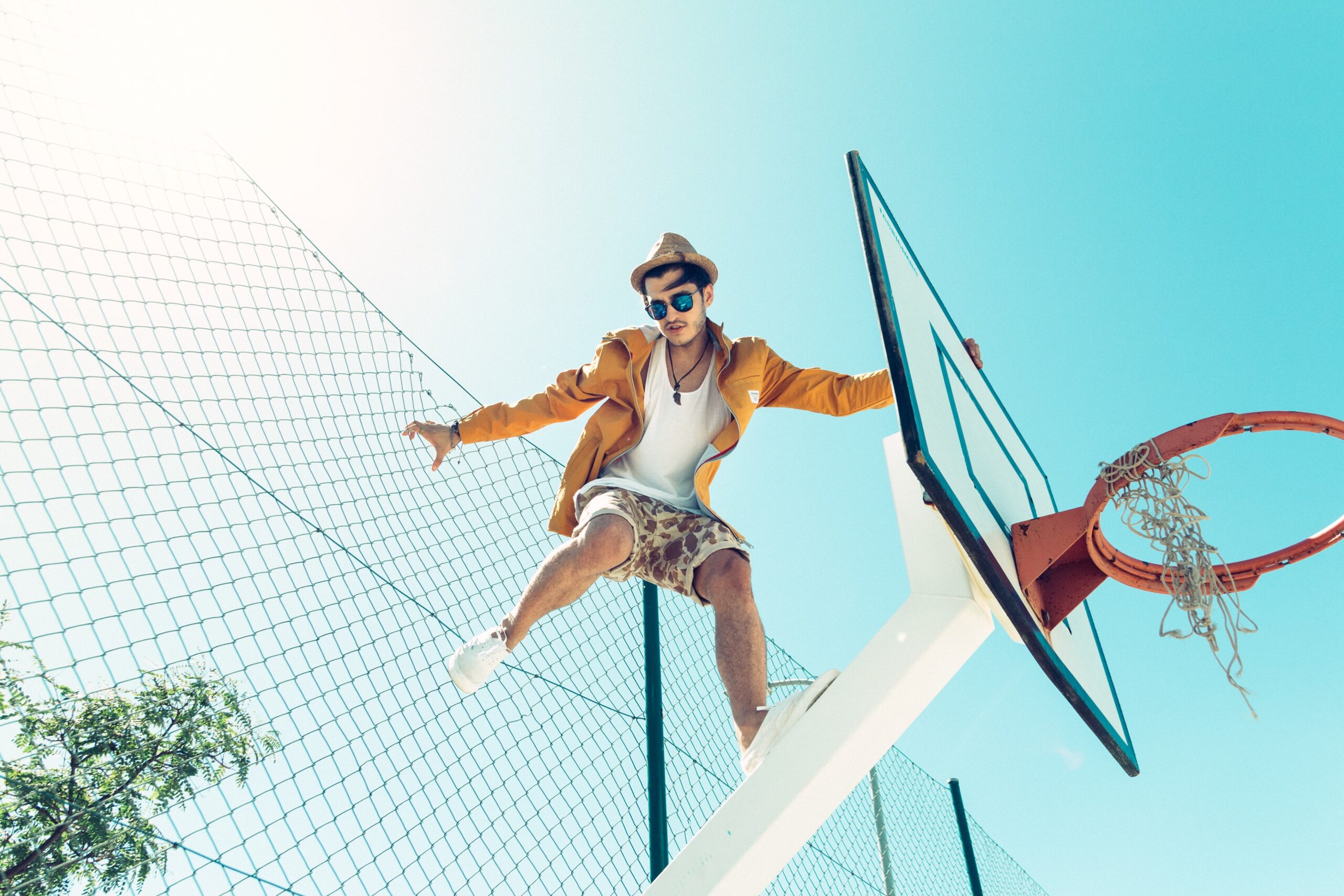 A young man in a yellow jacket, hat, and sunglasses sits on top of a basketball hoop, holding onto the backboard, with a chain-link fence and clear blue sky in the background.