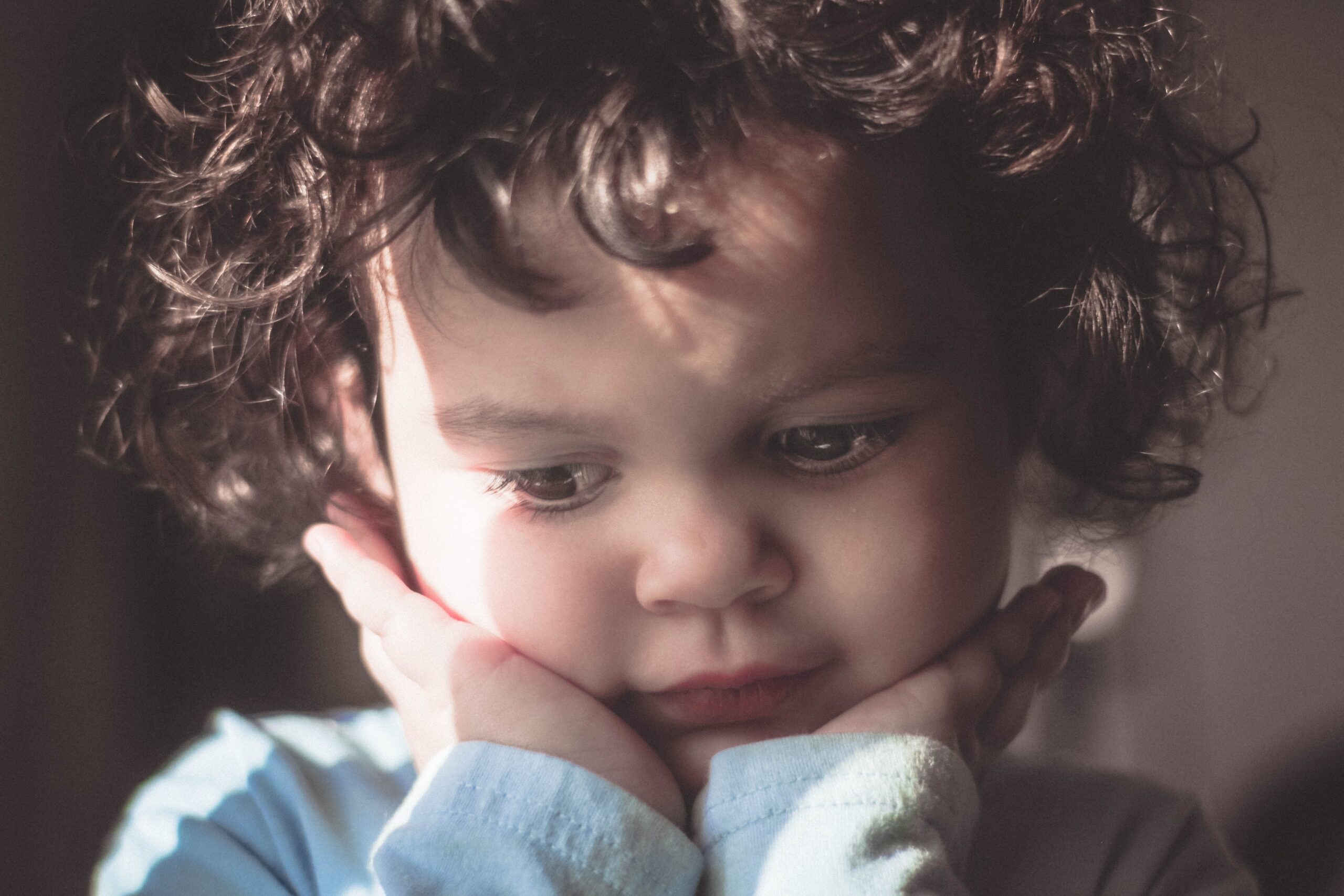 A young child with curly hair rests their chin on their hands, gazing downward with a thoughtful or pensive expression. Soft light highlights their face, emphasizing a calm and introspective mood.