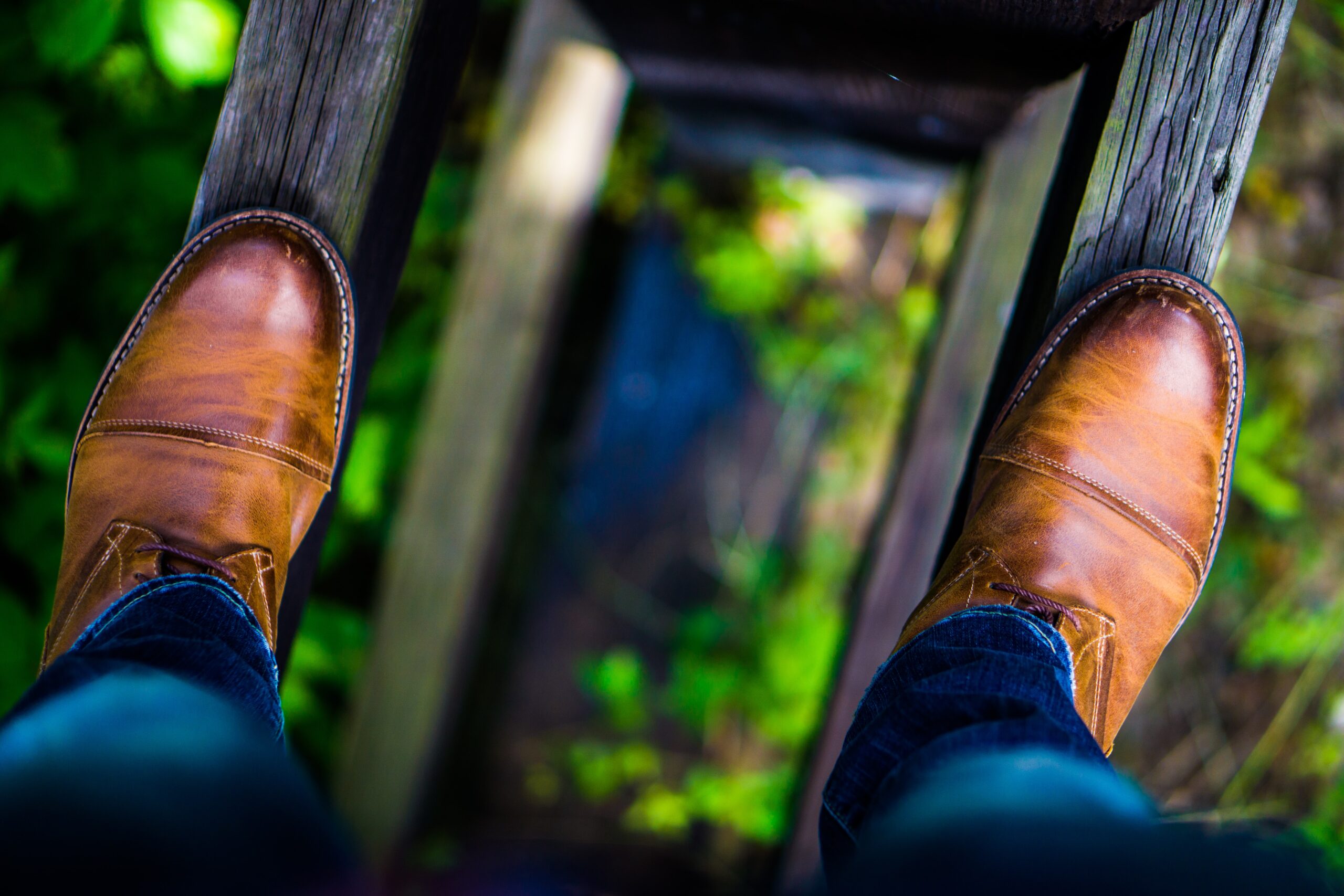 A person wearing brown leather shoes and jeans stands on a narrow wooden plank bridge, with green foliage visible below and around the planks. The scene is viewed from a first-person perspective.