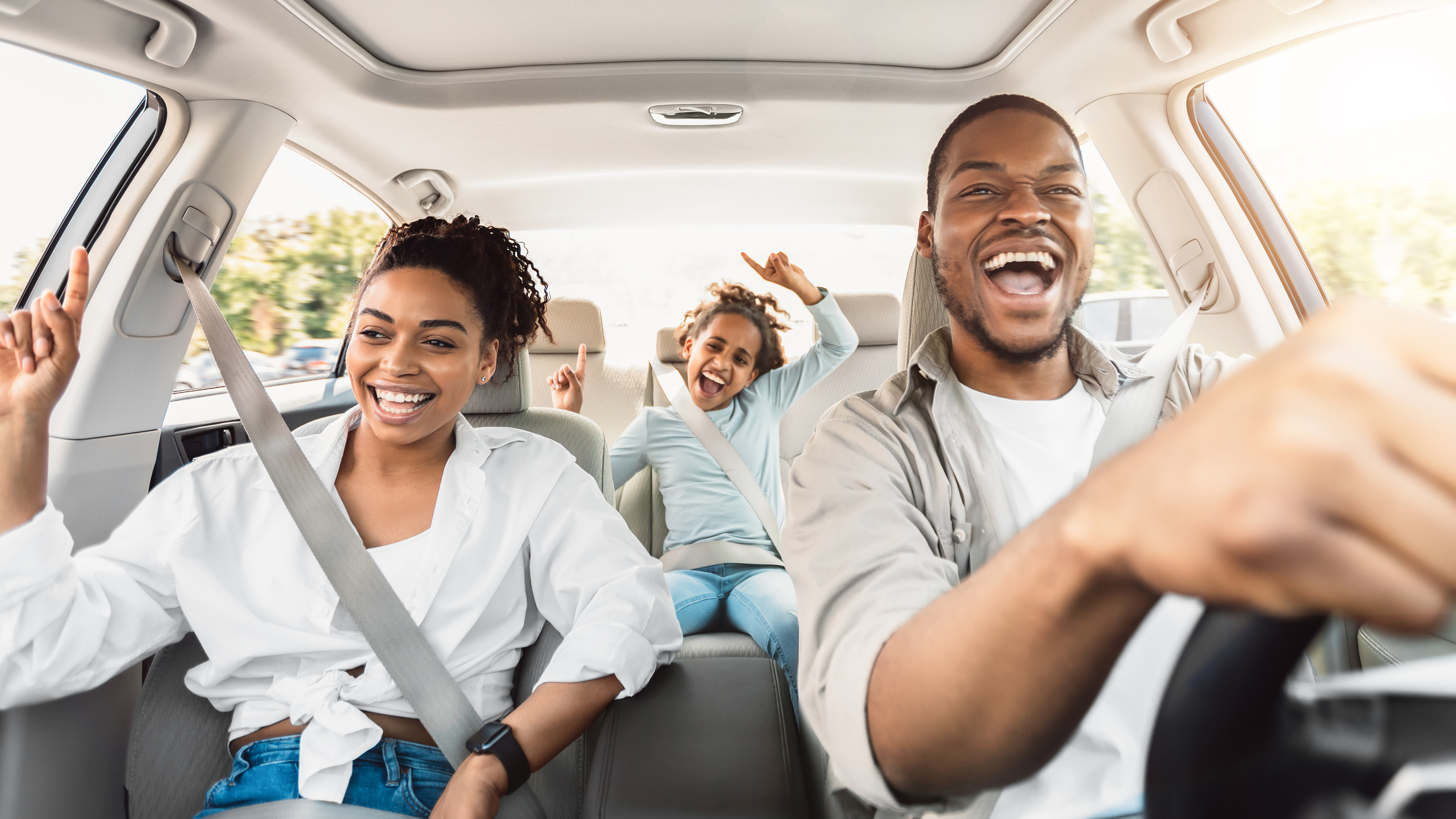 A smiling family of three, two adults in the front seats and a child in the back, enjoy a joyful car ride together on a sunny day. They are laughing and raising their hands, creating a happy, lively atmosphere.