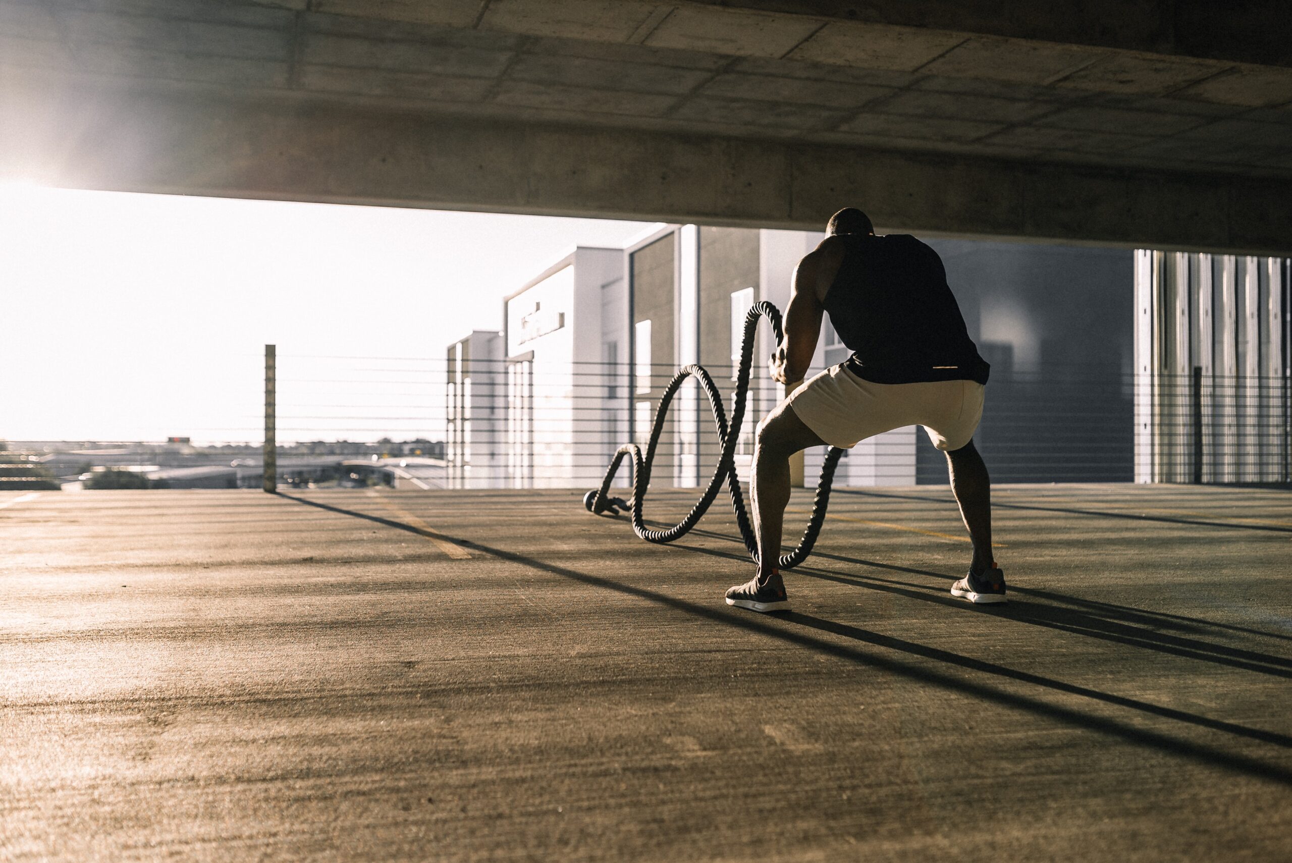 A person in athletic clothing exercises with battle ropes in an open, sunlit parking garage, with industrial buildings visible in the background.