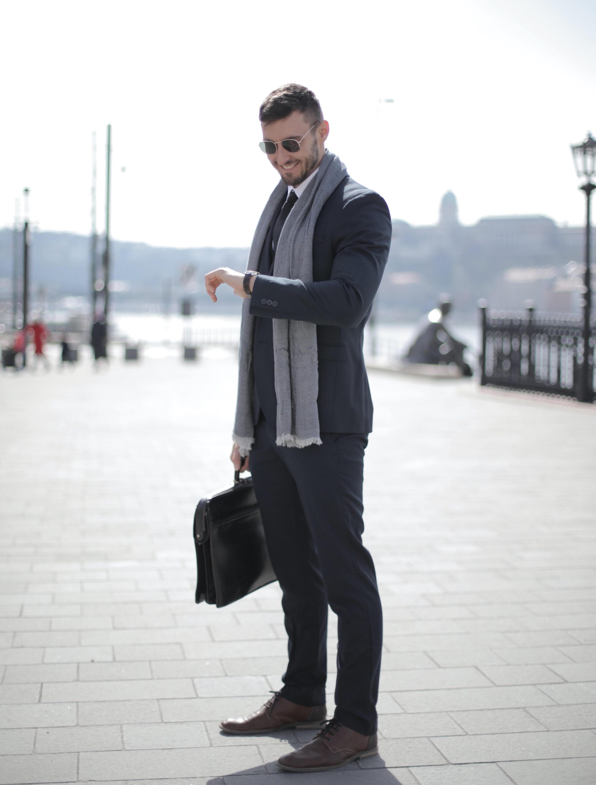 A man in a suit and scarf stands outdoors, holding a briefcase and looking at his watch. He is wearing sunglasses and smiling, with a cityscape and river in the background on a bright day.