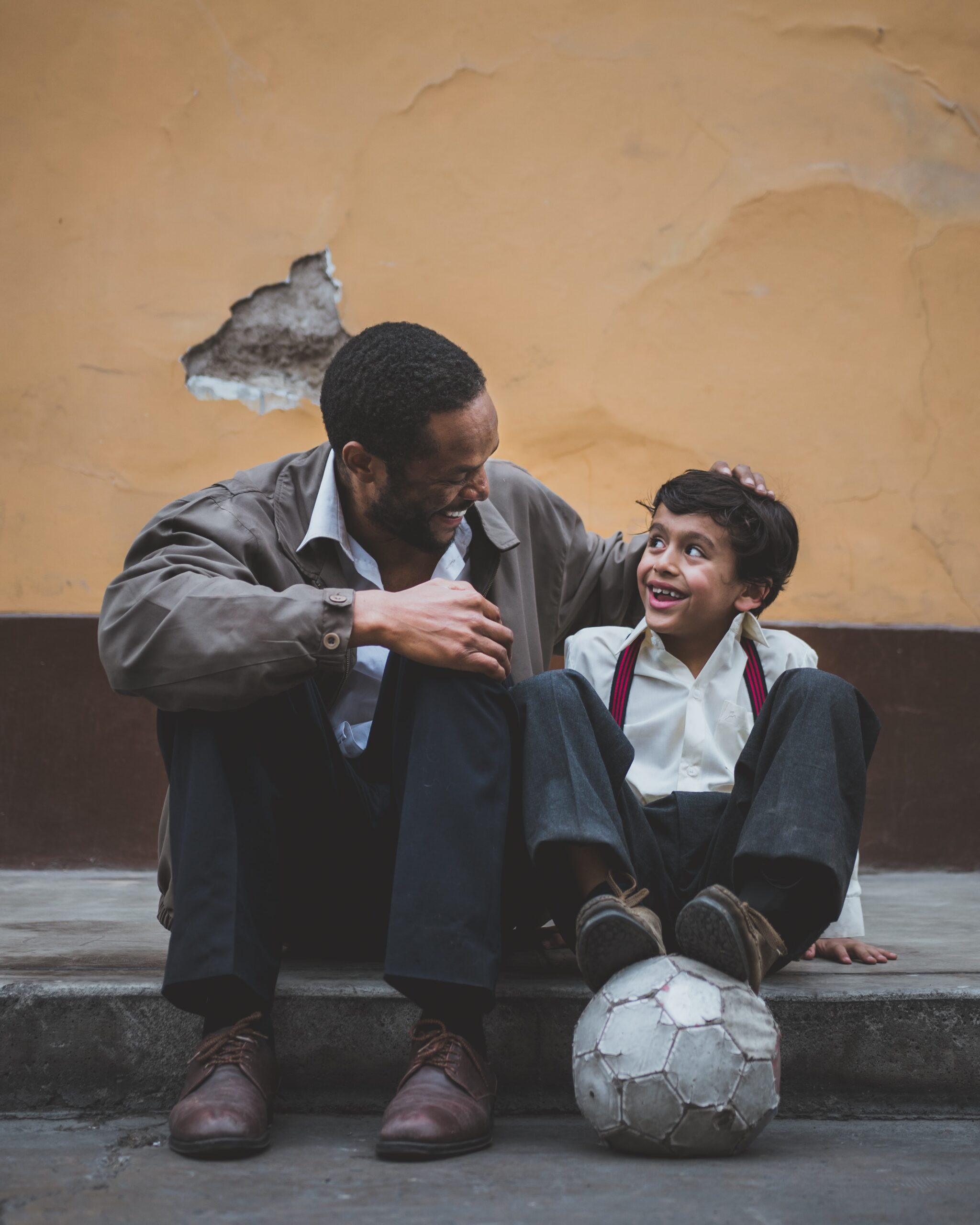 A man and a boy sit on a curb, smiling and laughing together. The man affectionately rests his hand on the boy’s head. A worn soccer ball is in front of them, and a weathered wall is in the background.
