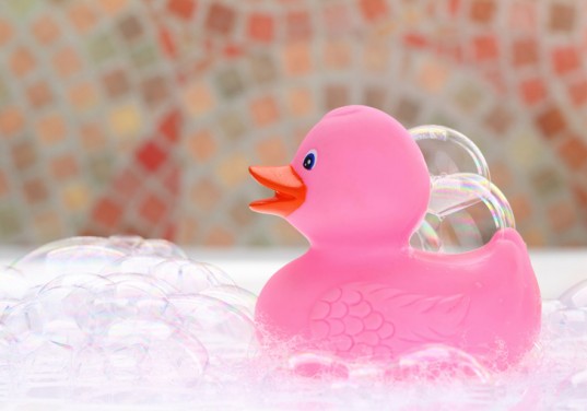 A pink rubber duck with an orange beak sits among bubbles, with a colorful, out-of-focus mosaic background.