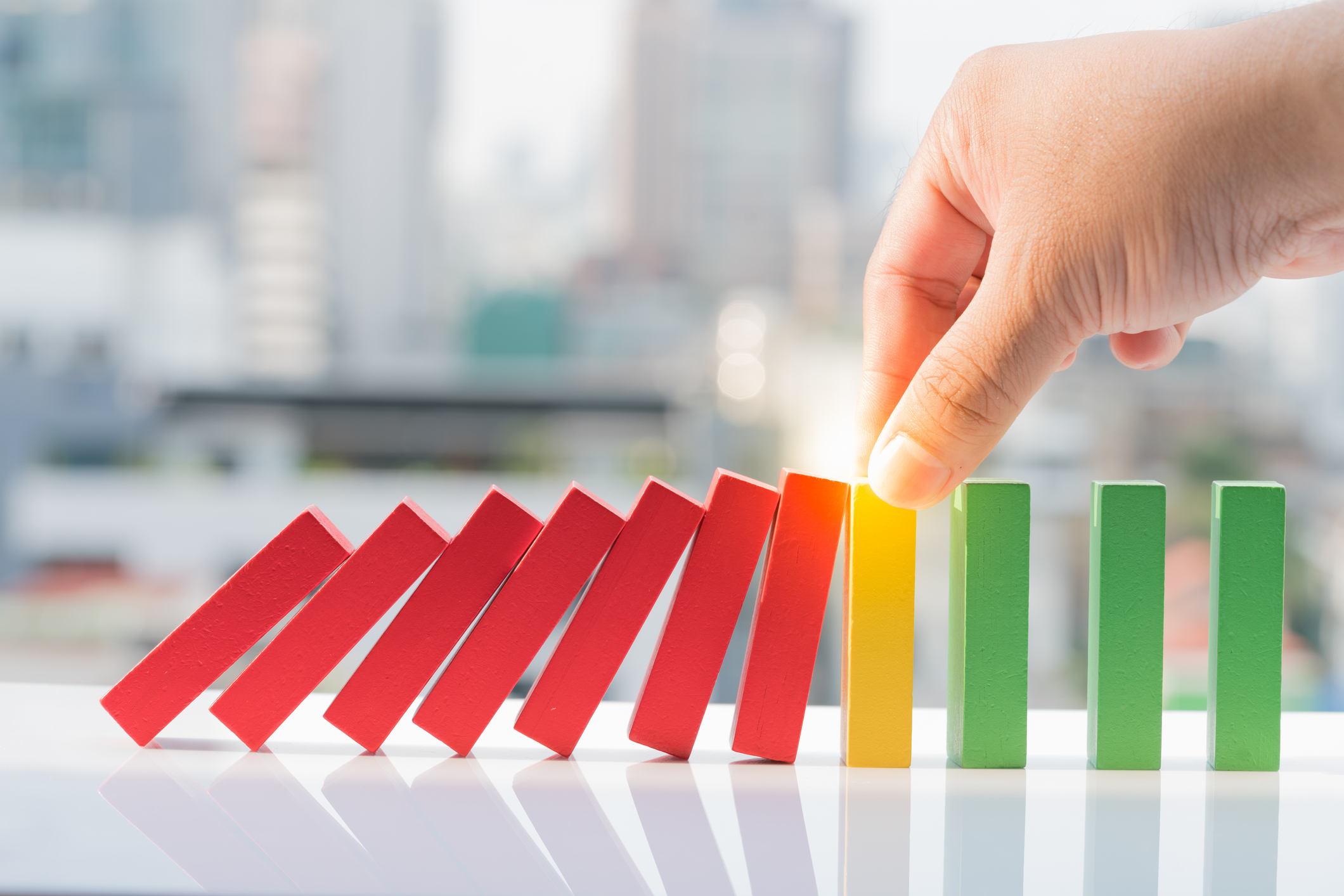 A hand stops a row of falling red dominoes from knocking over upright yellow and green dominoes, with a blurred cityscape in the background.