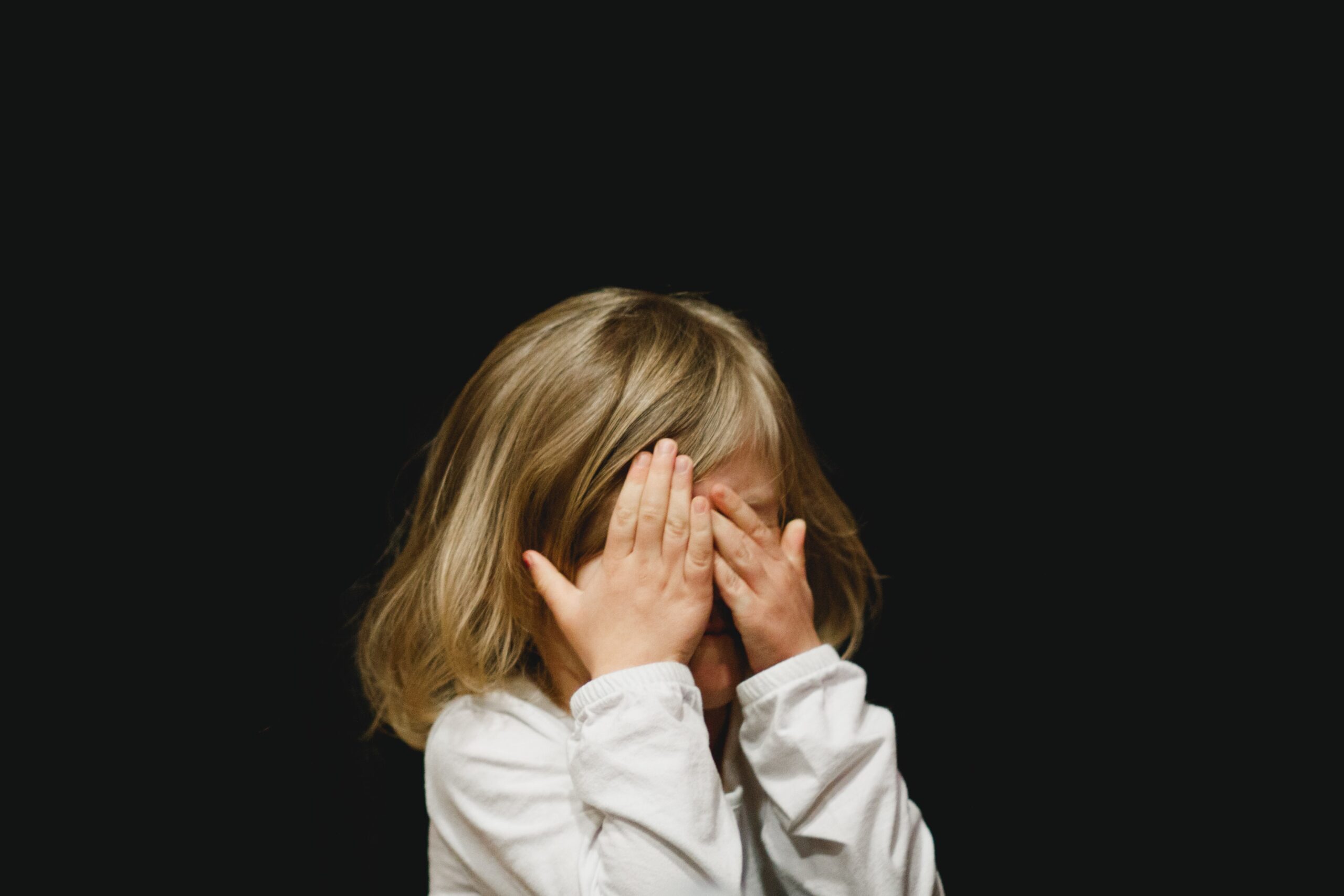 A child with shoulder-length blonde hair wearing a white shirt covers their face with both hands against a plain black background.