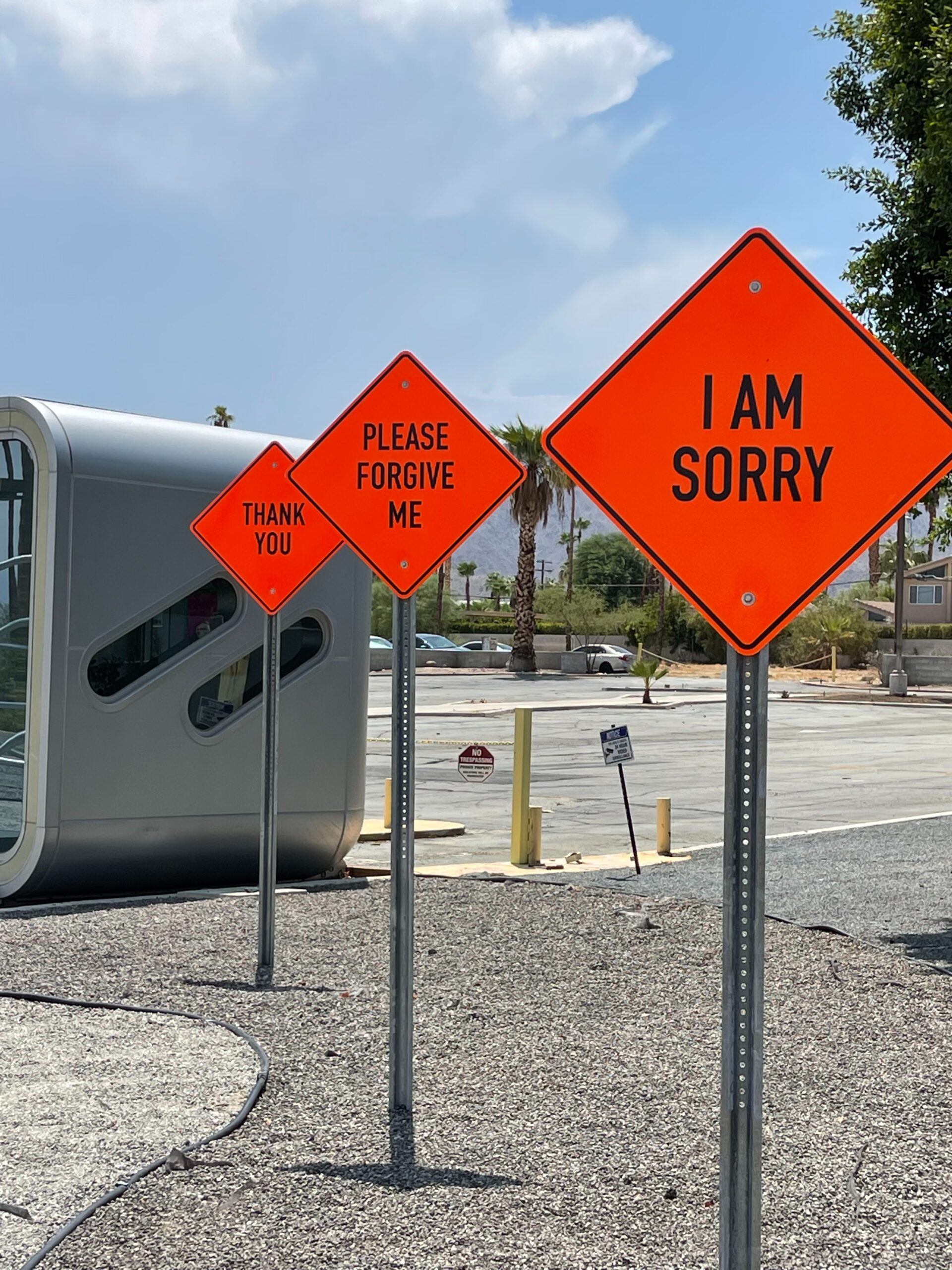 Three orange diamond-shaped road signs read THANK YOU, PLEASE FORGIVE ME, and I AM SORRY. They stand in a row beside a modern building and an empty parking lot on a sunny day.