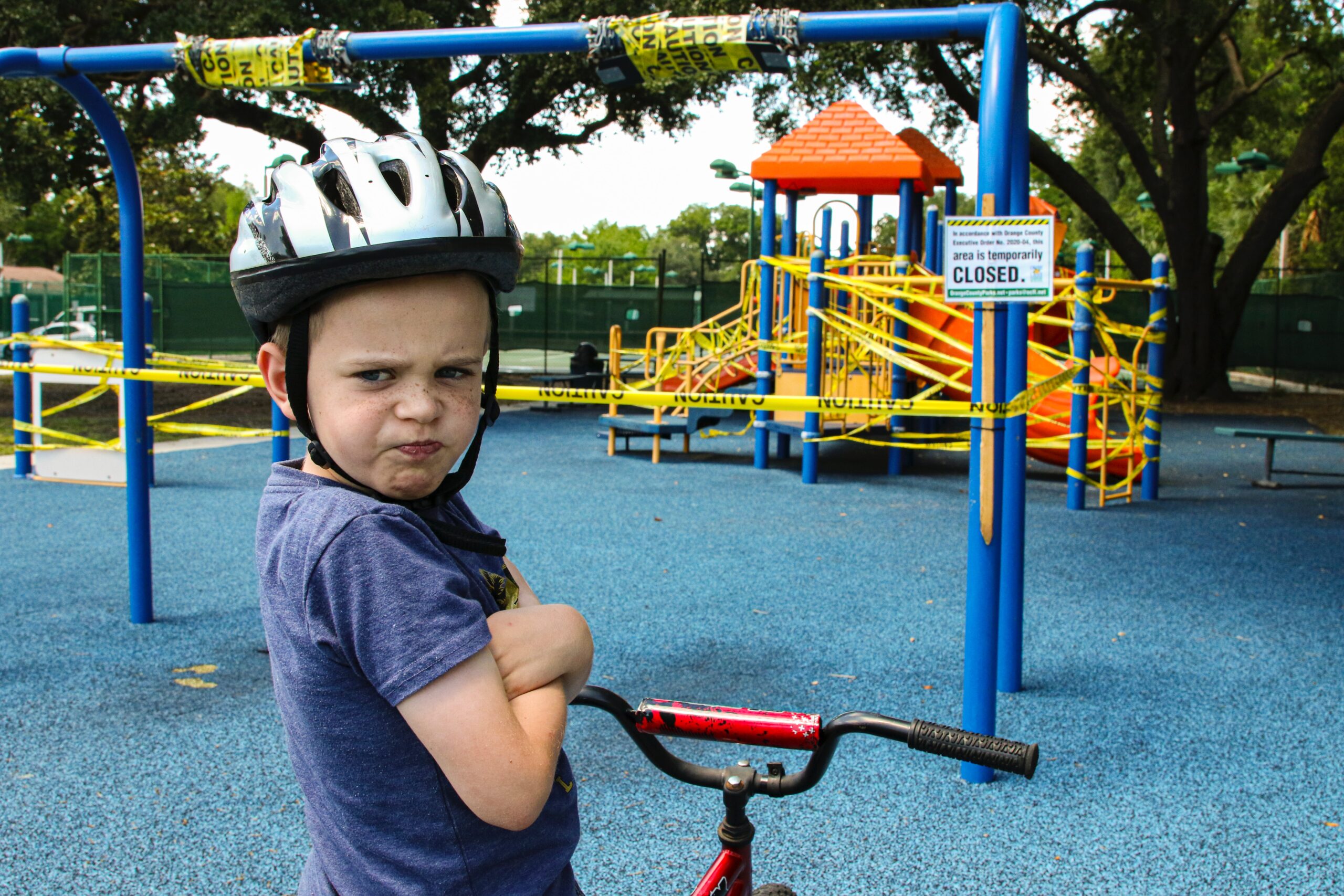 A young boy wearing a bike helmet pouts while holding his bike at a playground closed off with caution tape and a “closed” sign, learning firsthand about the kind vs. nice difference as he faces the restrictions. The playground equipment is visible in the background.