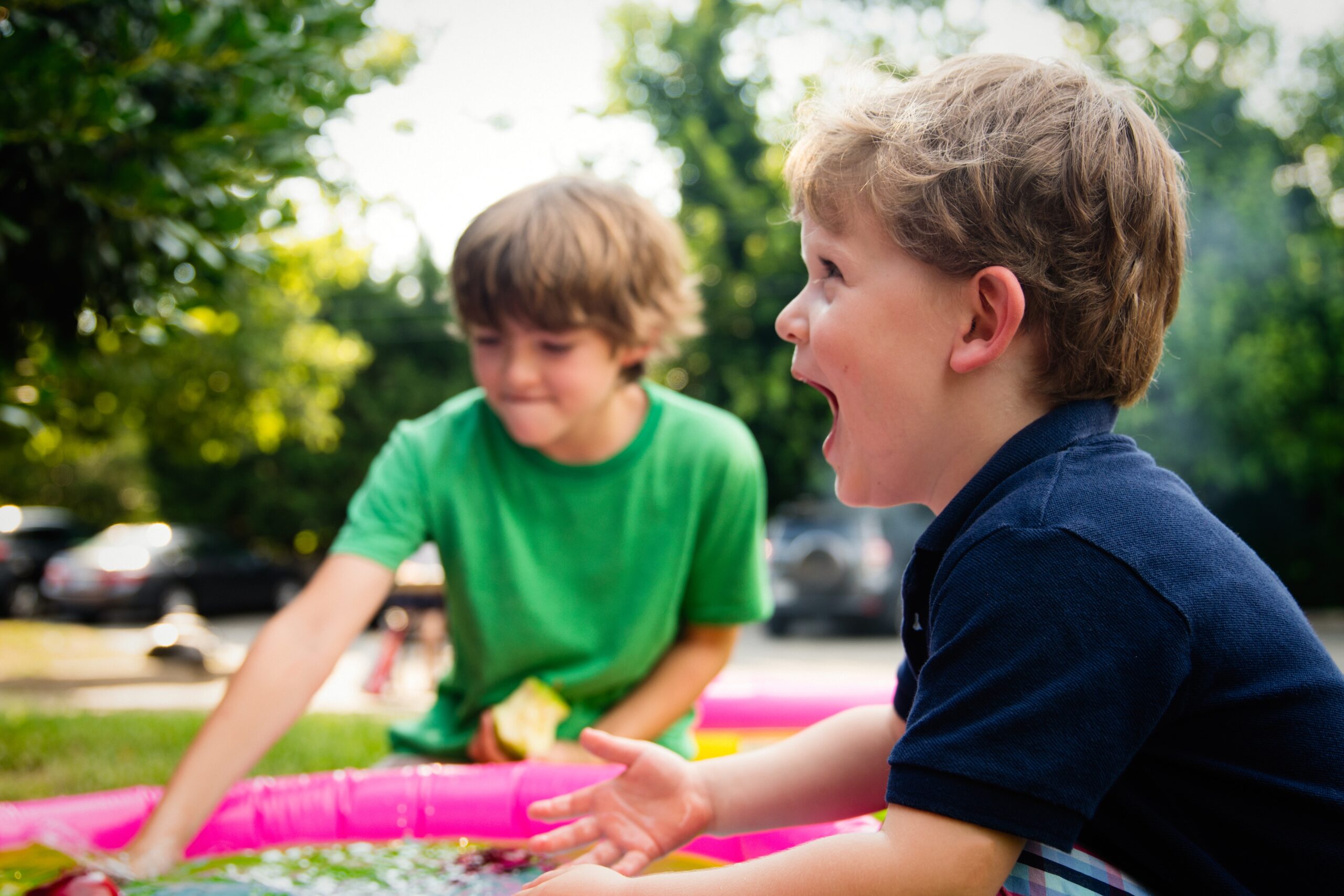 Two young boys play outside next to a pink inflatable pool. Amid fatherhood challenges and ego, one boy in a green shirt reaches toward the water while the other, in a navy shirt, laughs with excitement. Trees and parked cars fill the background.