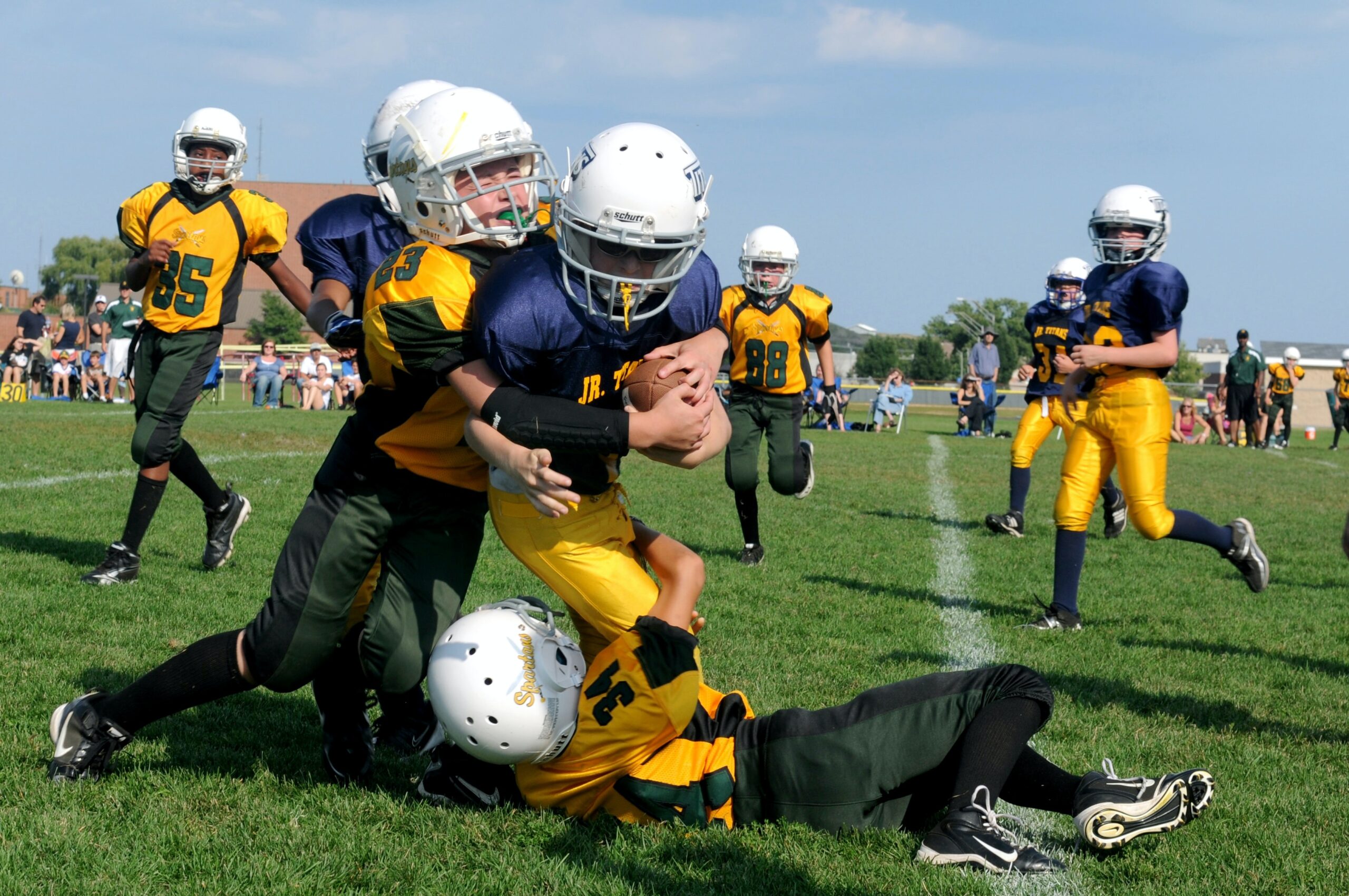 Youth football players in helmets and uniforms tackle a player holding the ball during a game on a grassy field, breaking gender stereotypes, as other players and spectators watch in the background.