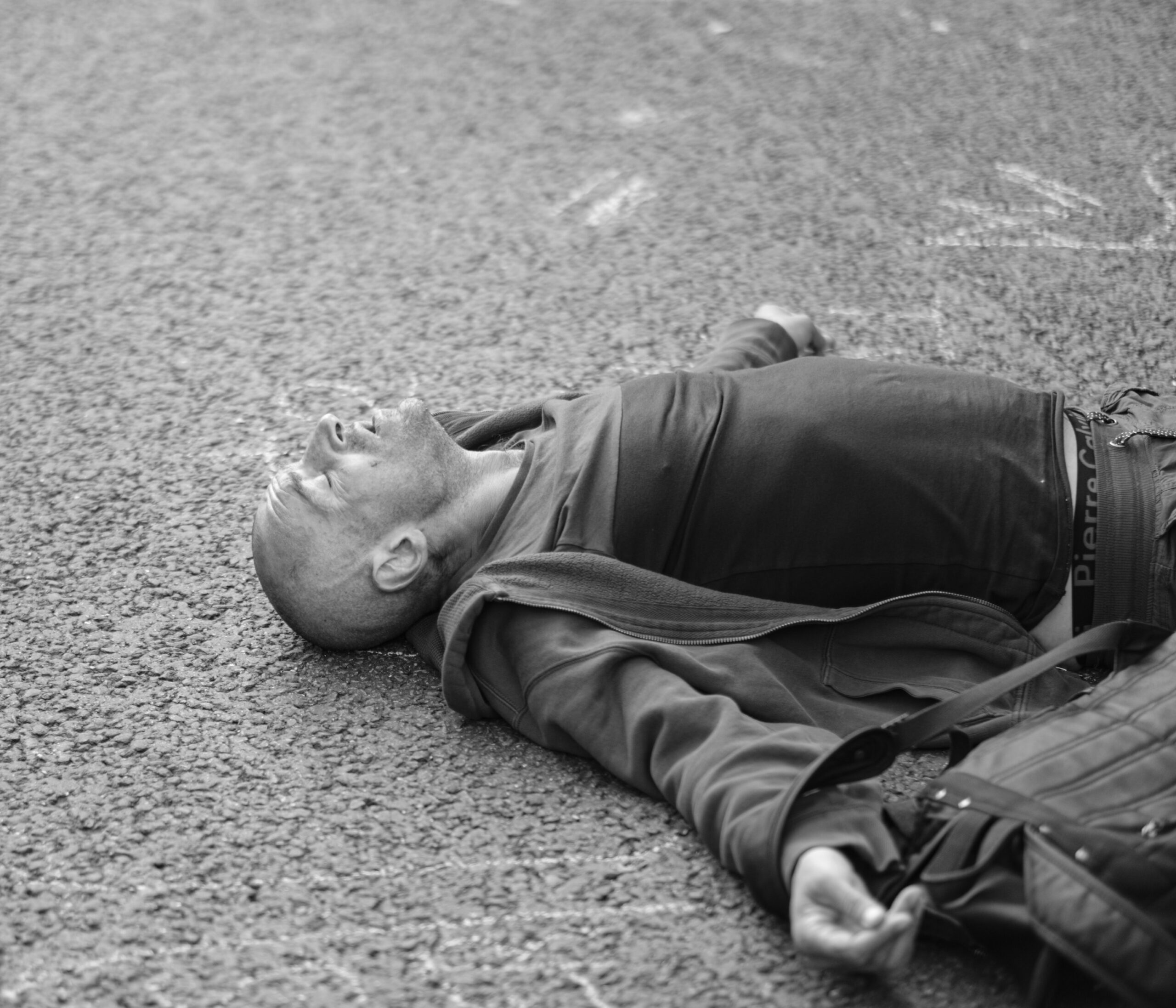 A man lies on his back on a rough asphalt surface with his arms spread out, eyes closed, mouth slightly open, as if he chose to quit parenting strategically. He wears a jacket, t-shirt, and bag across his shoulder. The image is in black and white.